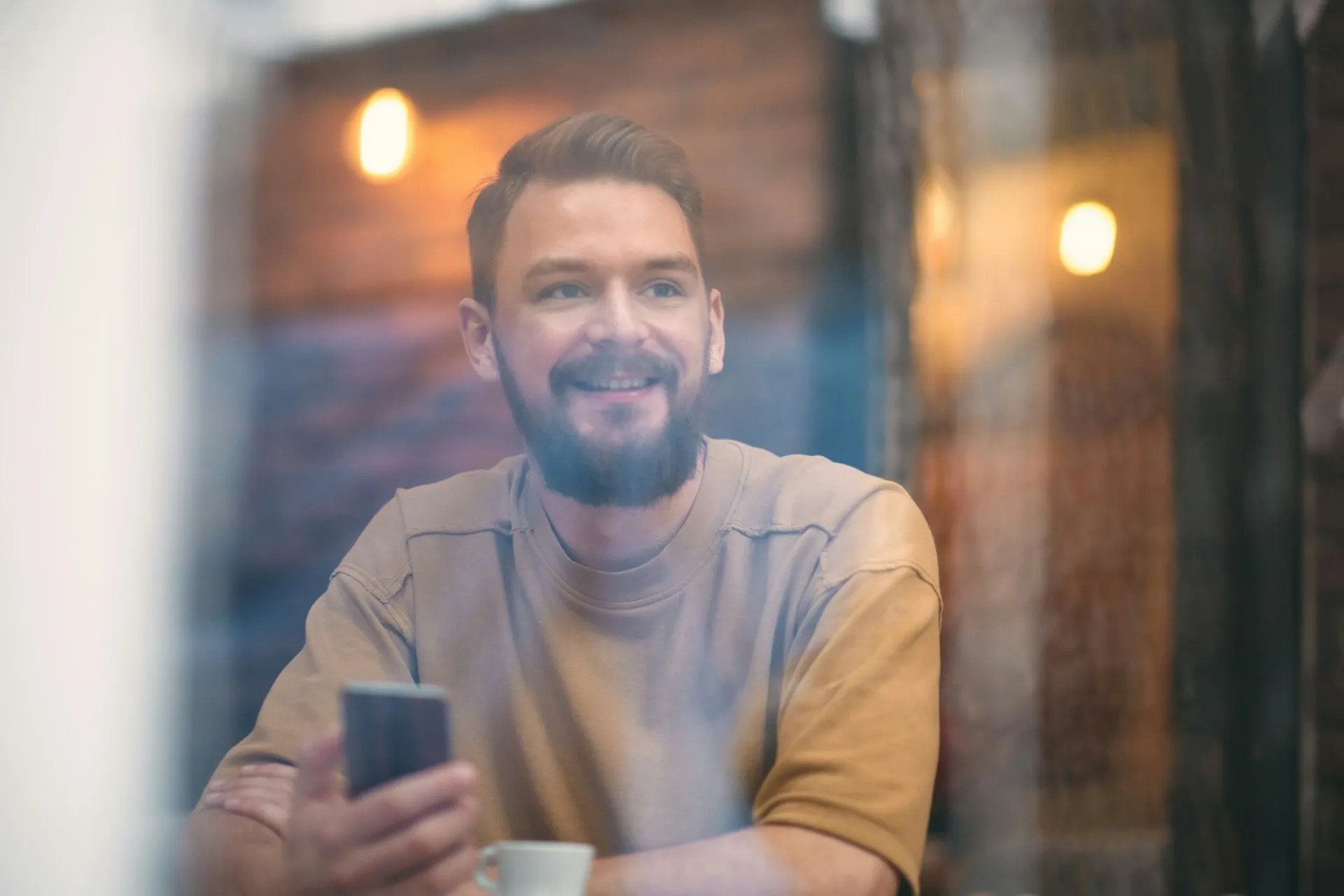 Smiling man holding a smartphone and sitting at a table with a coffee cup indoors.