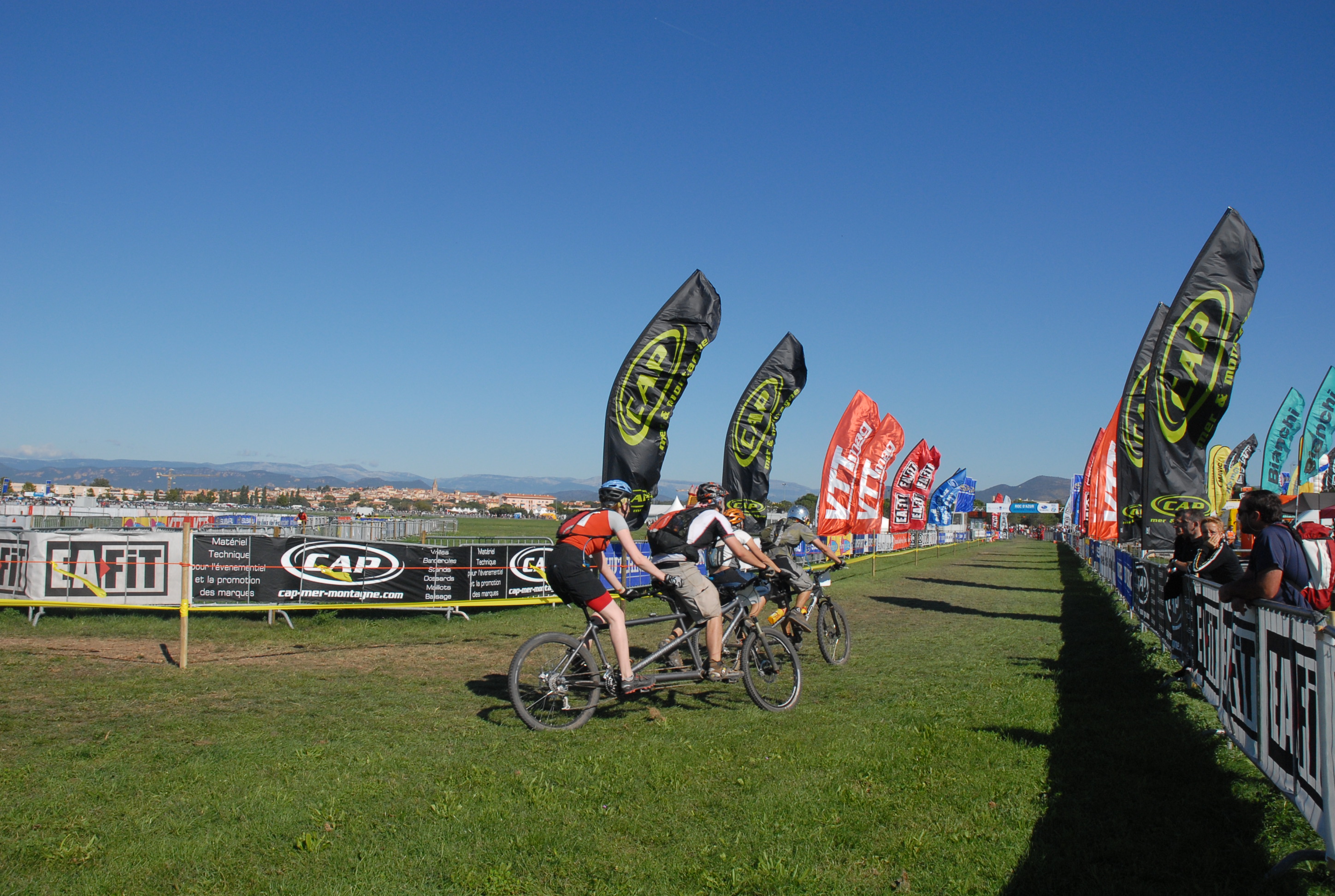 course de tandem VTT avec plusieurs coureurs au départ sur un événement sportif entouré de drapeaux promotionnels