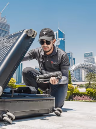 Man wearing sunglasses and a cap crouching on a city street, unpacking a drone from a large black rolling case with modern skyscrapers and palm trees in the background.