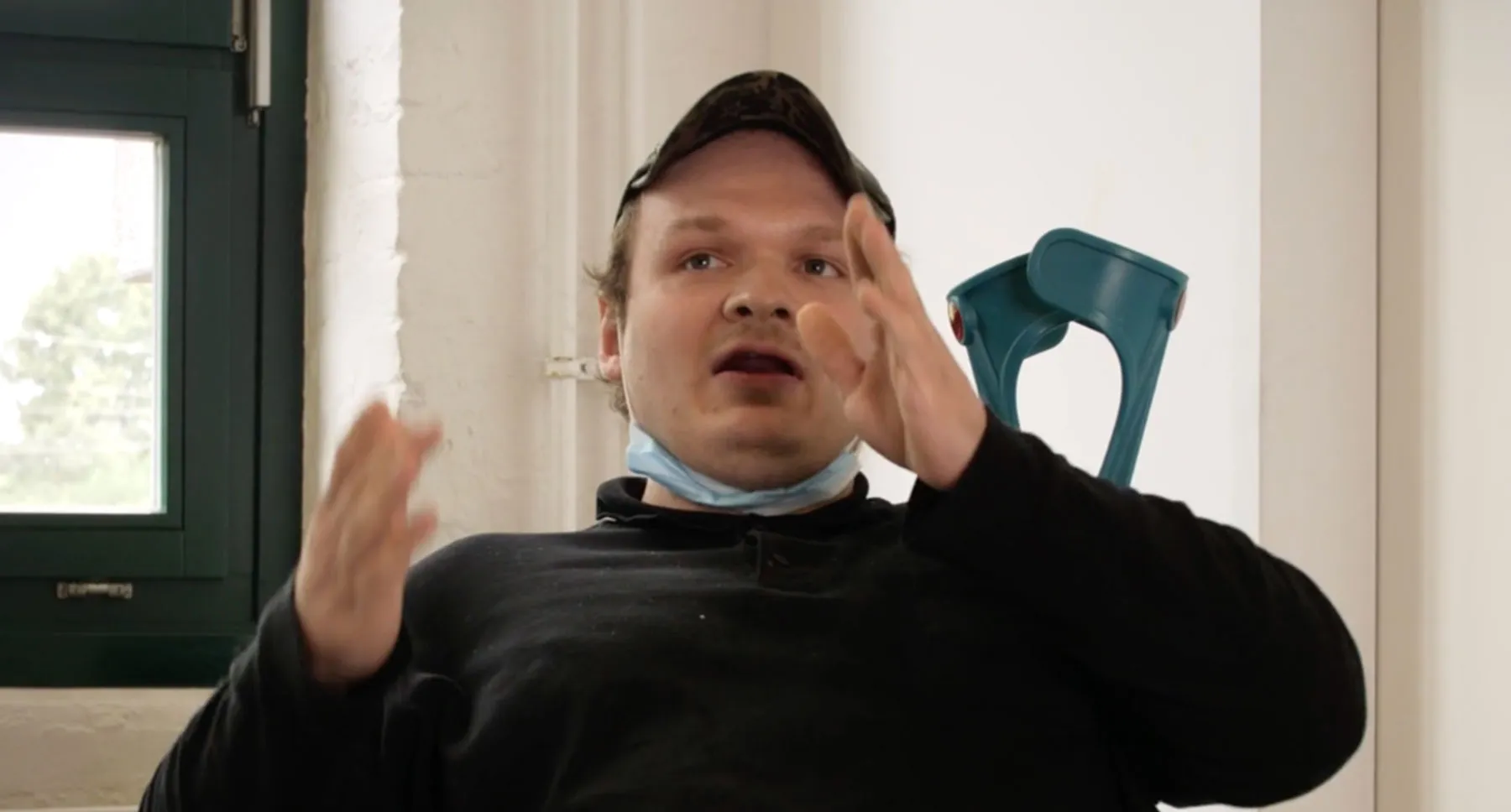 Ein Mann gestikuliert mit den Händen. Hinter ihm sind zwei Gehstützen.