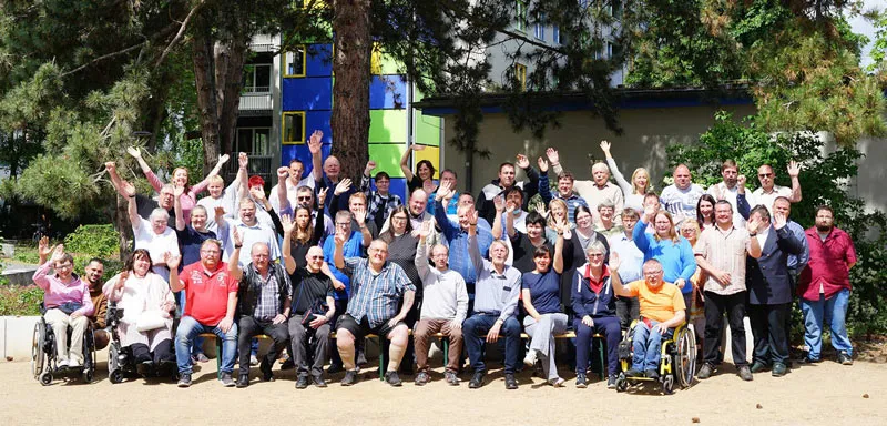 Mitglieder der Werkstatträte Deutschland winken beim Gruppenfoto in die Kamera. Im Hintergrund Bäume und ein buntes Gebäude.