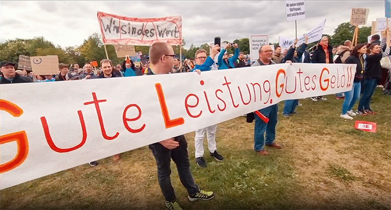 Menschen bei einer Demonstration im Freien halten ein großes Banner mit der Aufschrift „Gute Leistung, Gutes Geld!" Im Hintergrund weitere Teilnehmende mit Schildern.