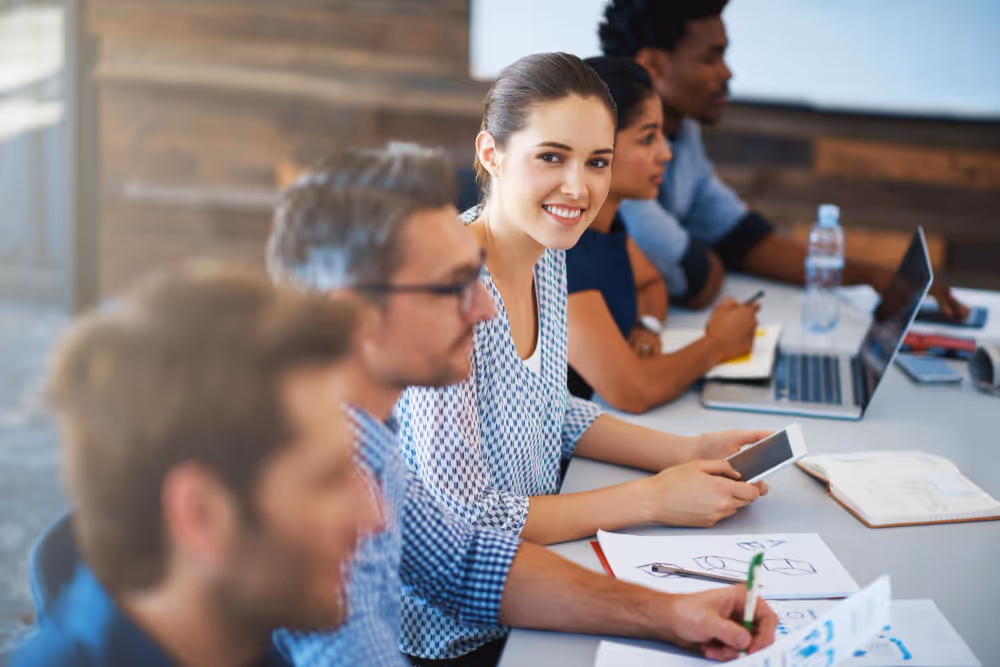 Fünf junge Erwachsene sitzen an einem Tisch und arbeiten gemeinsam, eine Frau lächelt in die Kamera.