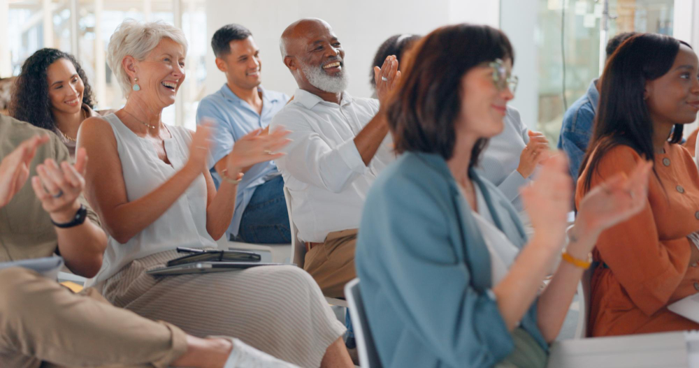 Eine vielfältige Gruppe von Erwachsenen sitzt in einem Raum und klatscht begeistert bei einer Veranstaltung.
