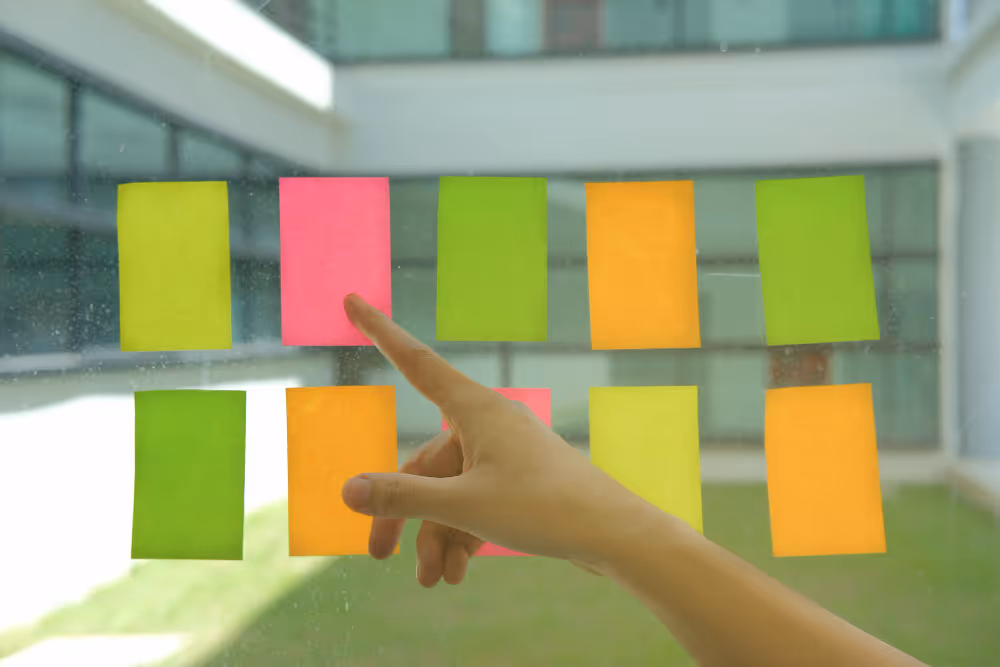 Hand zeigt auf eine rosa Haftnotiz unter mehreren bunten Haftnotizen an einem Fenster.