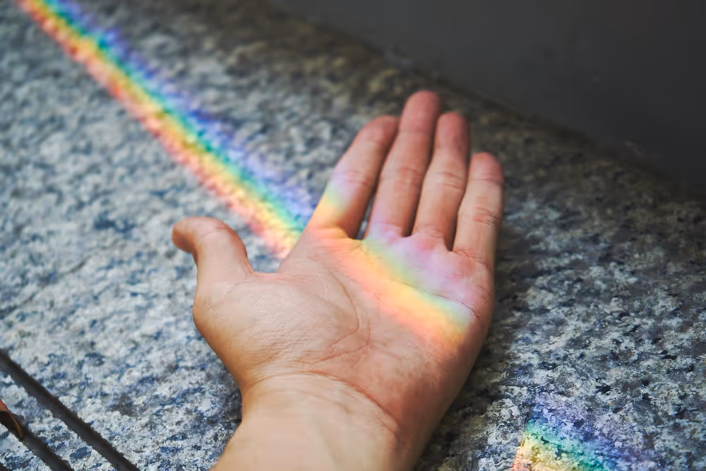 Hand mit ausgestreckter Handfläche, auf der ein Regenbogenlichtstreifen auf grauem Steinboden fällt.