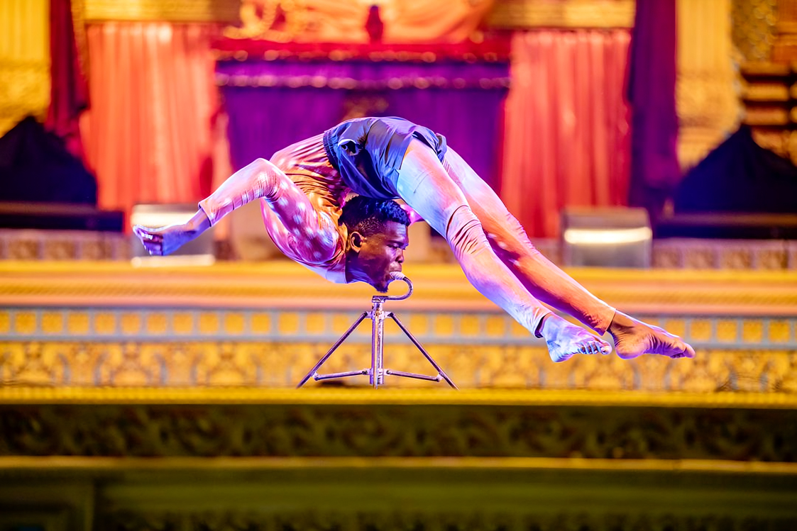 Contortionist balancing on a small metal stand with his mouth, arching his body backwards in an ornate theater setting.