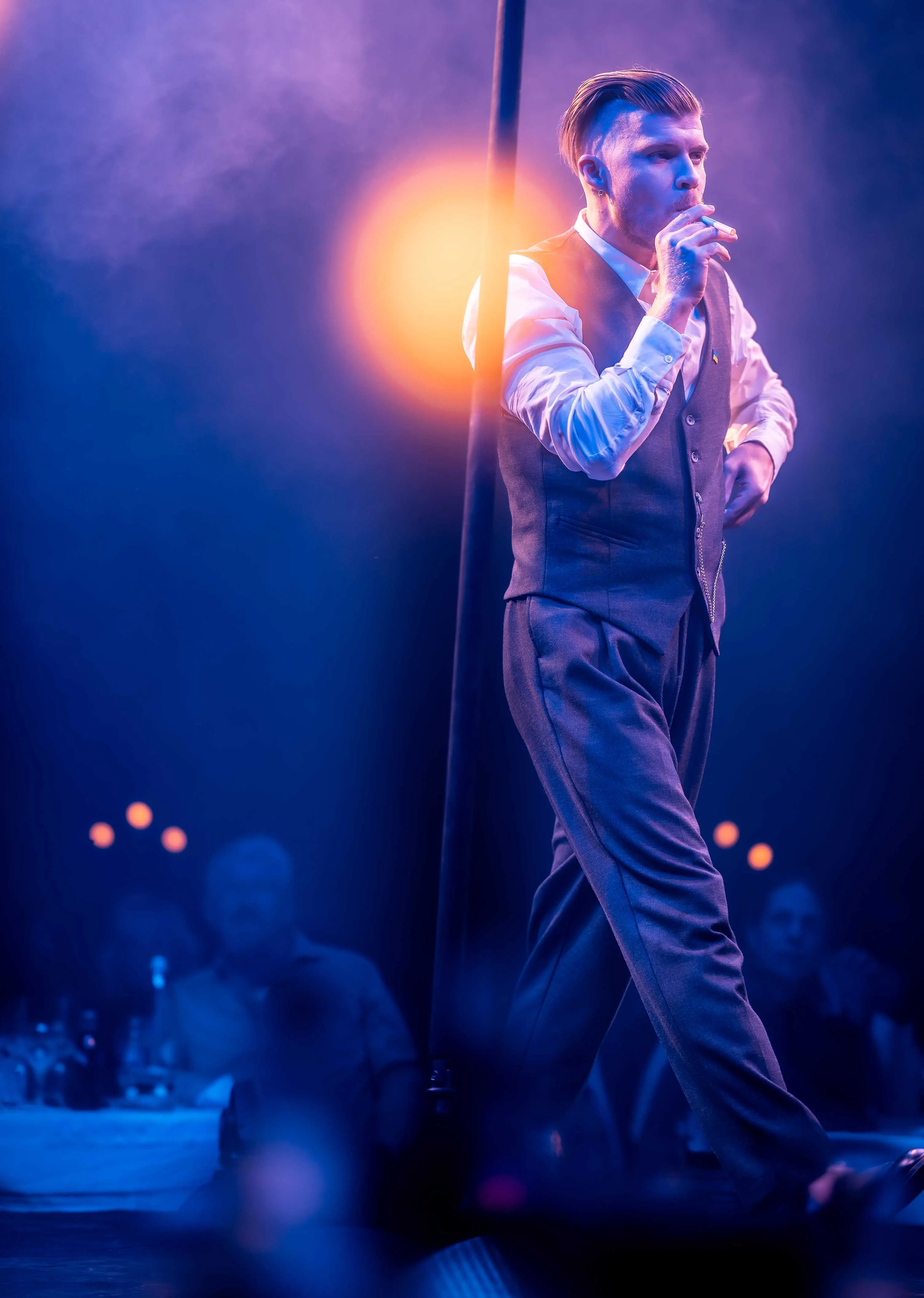 Man in vintage formal attire smoking a cigarette on stage with blue and orange lighting.