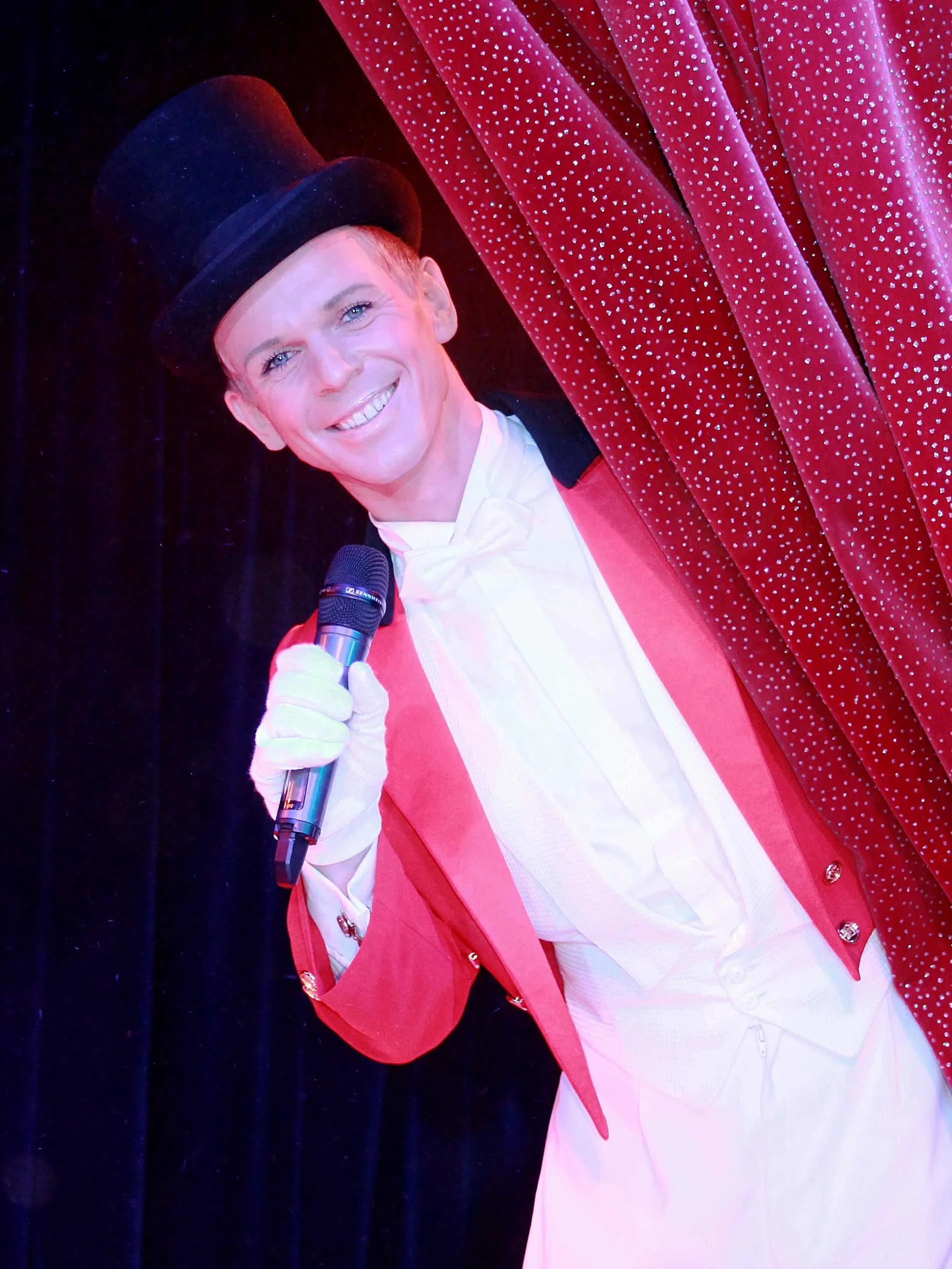 Smiling performer in a red tailcoat, white gloves, and top hat holding a microphone and peeking from behind a red curtain with white dots.