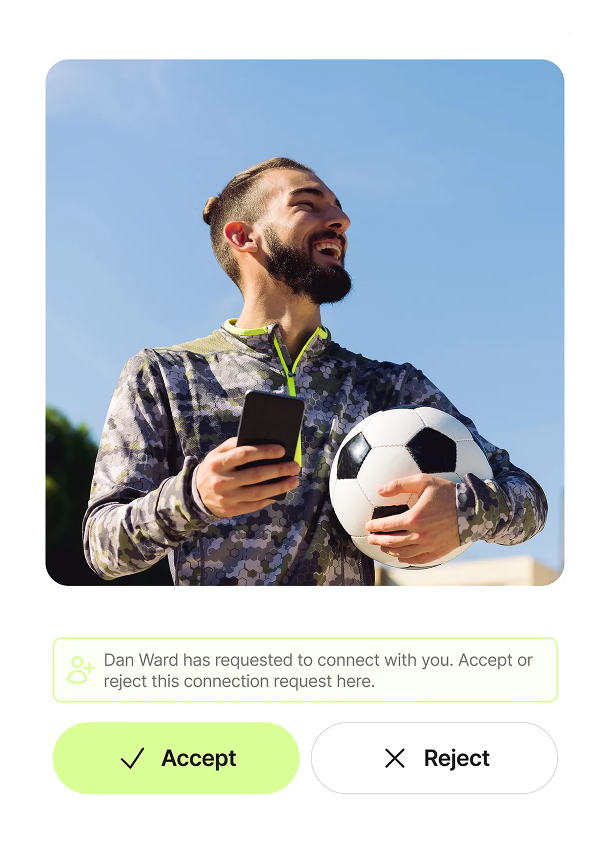 Smiling man holding a soccer ball in one arm and a smartphone in the other, set against a clear blue sky.