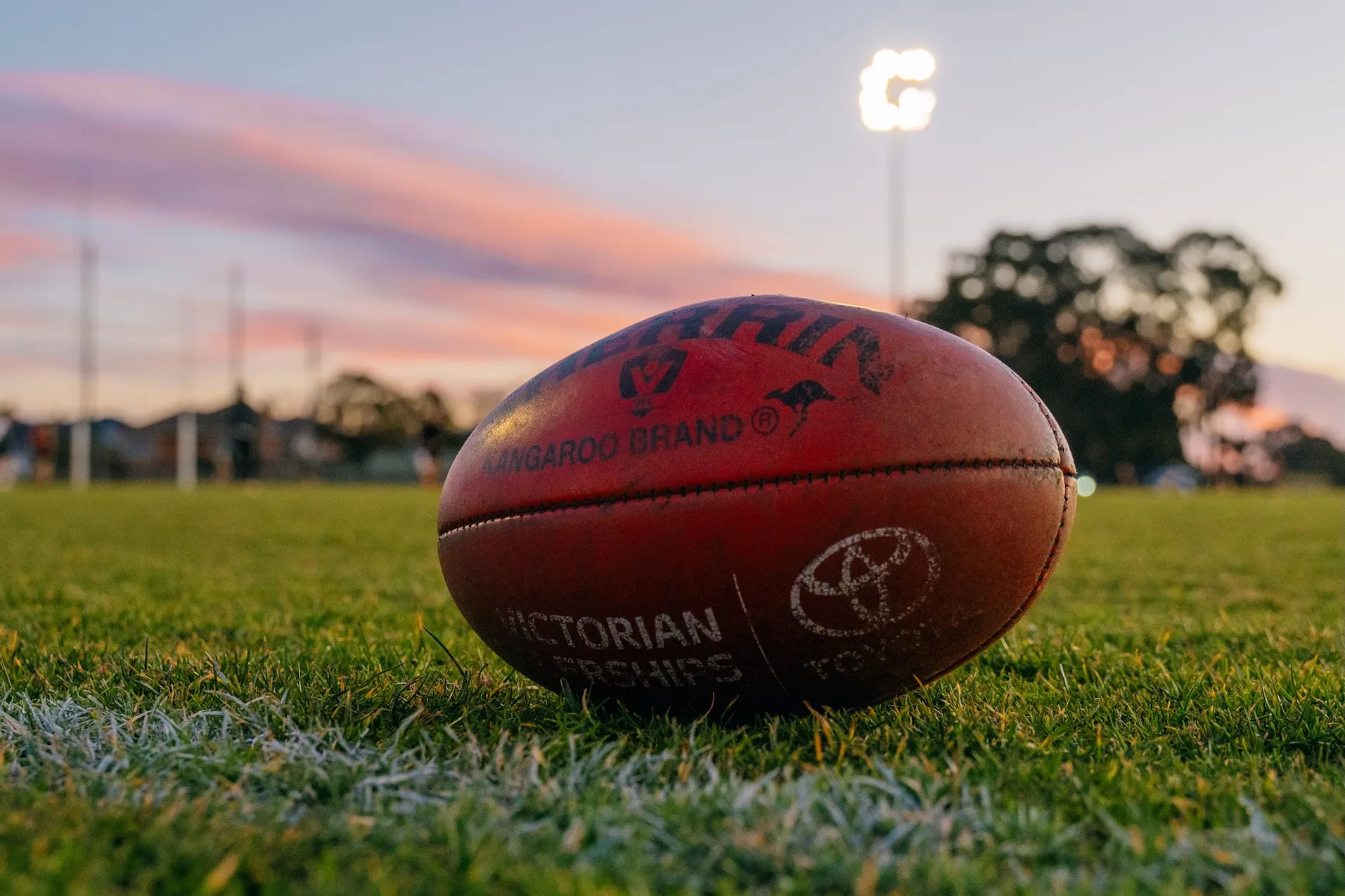 Close-up of an Australian rules football on grass with goal posts and evening sky in the background.