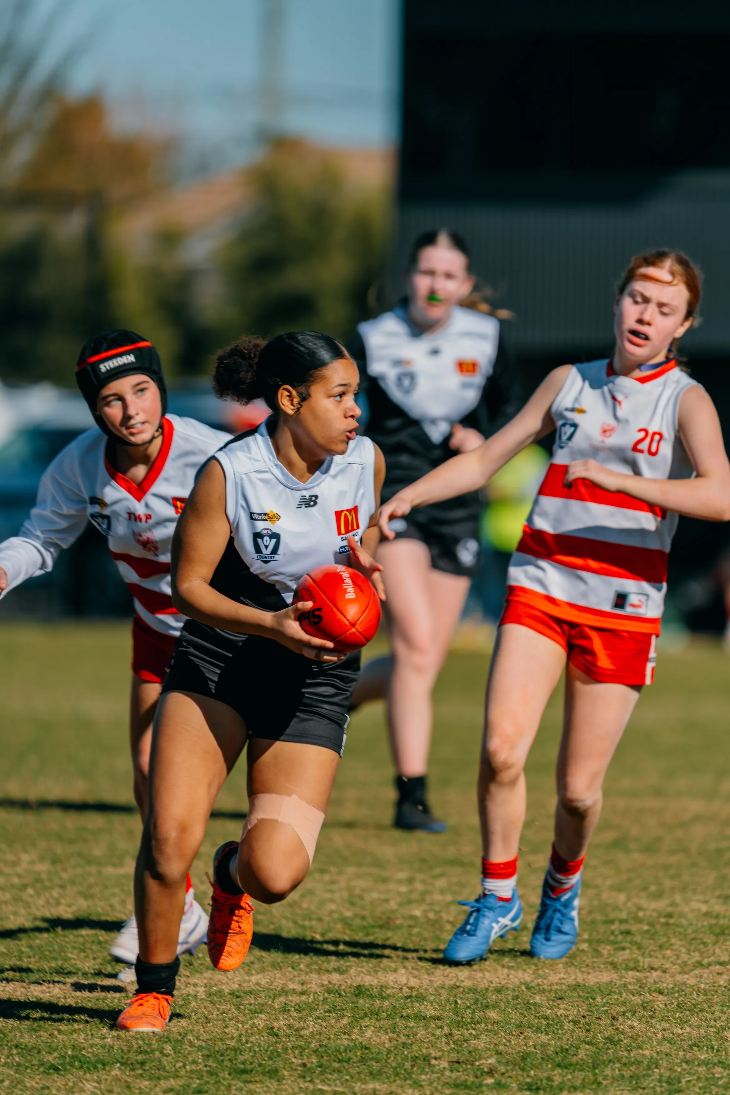 Female Australian rules football player in black and white uniform runs with the ball while being pursued by opponents in red and white uniforms.