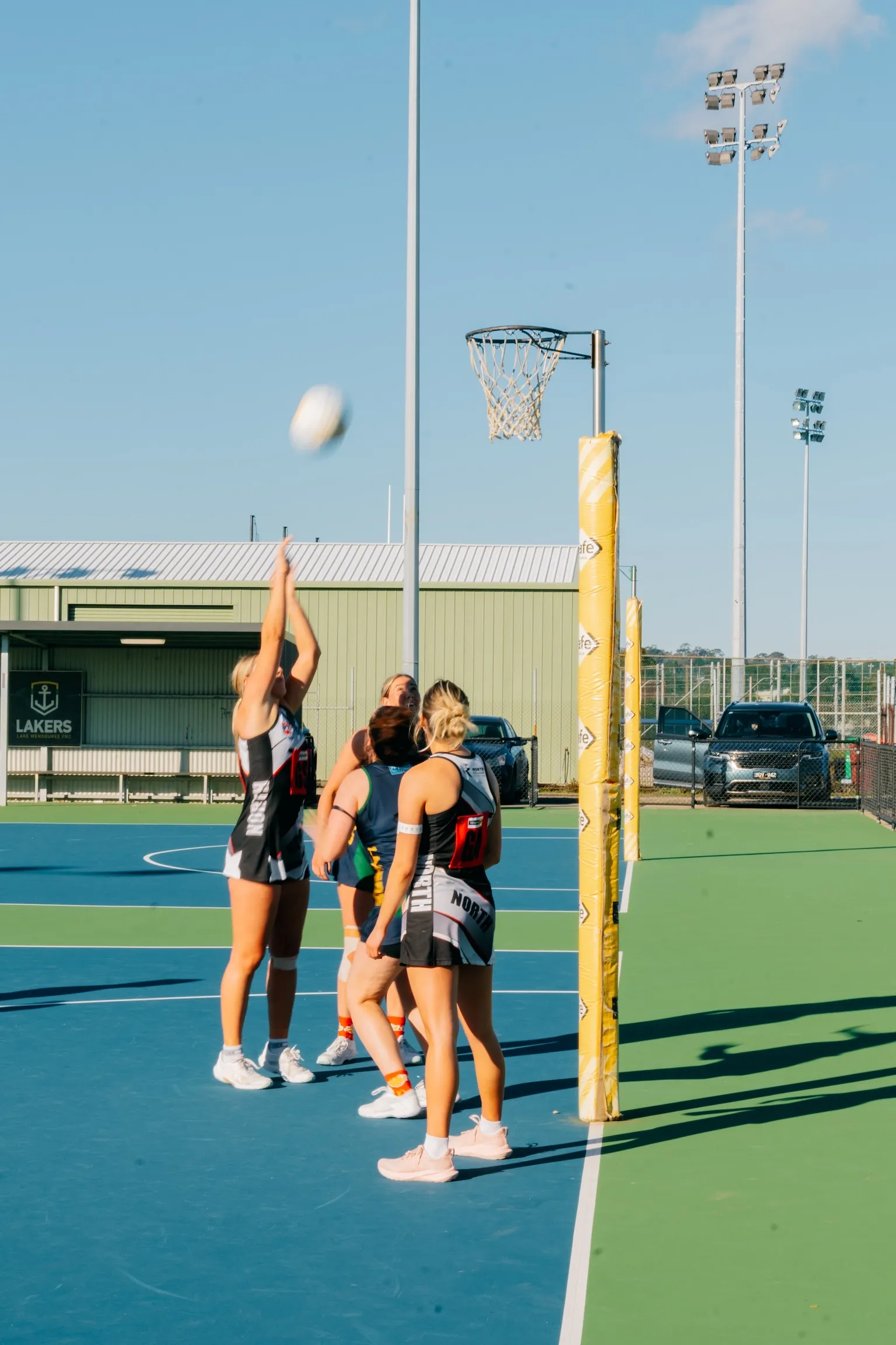 Female netball players in black and white uniforms play on an outdoor court under clear blue sky, with one player shooting the ball towards the hoop.
