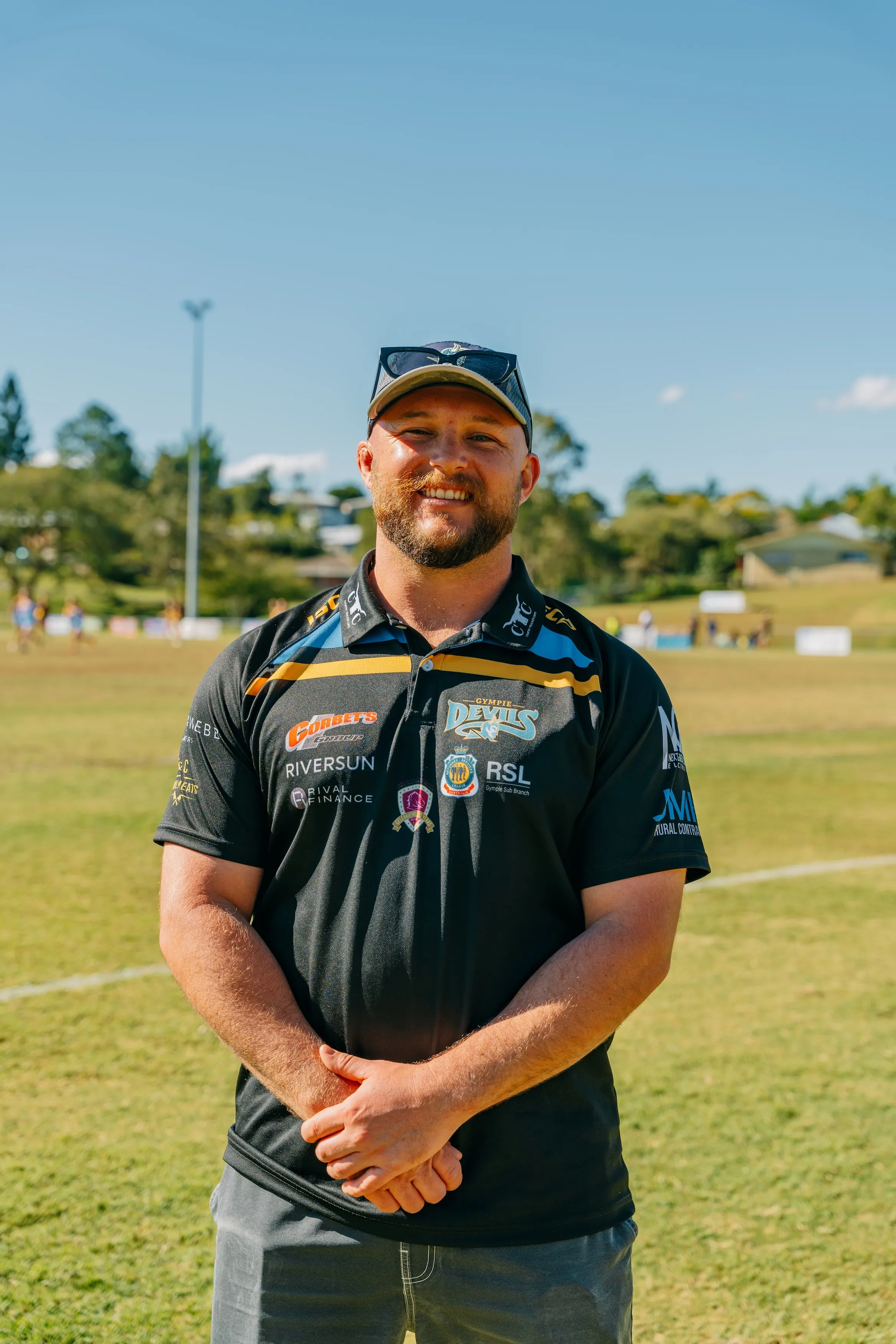Smiling man wearing a Gympie Devils sports polo and cap standing on a grassy sports field.