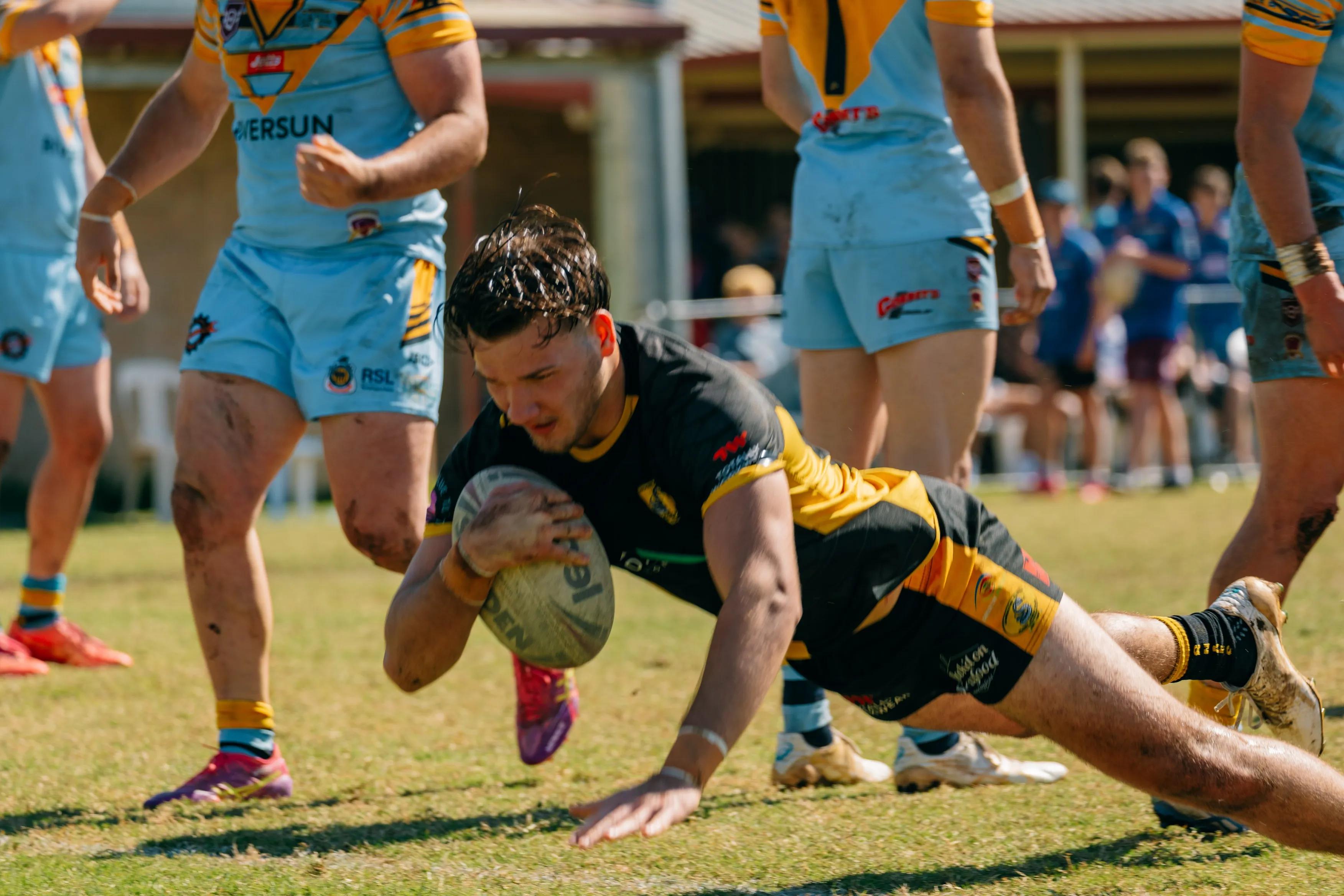 Rugby player in black and yellow uniform dives to ground holding ball during a match with opponents in light blue uniforms nearby.