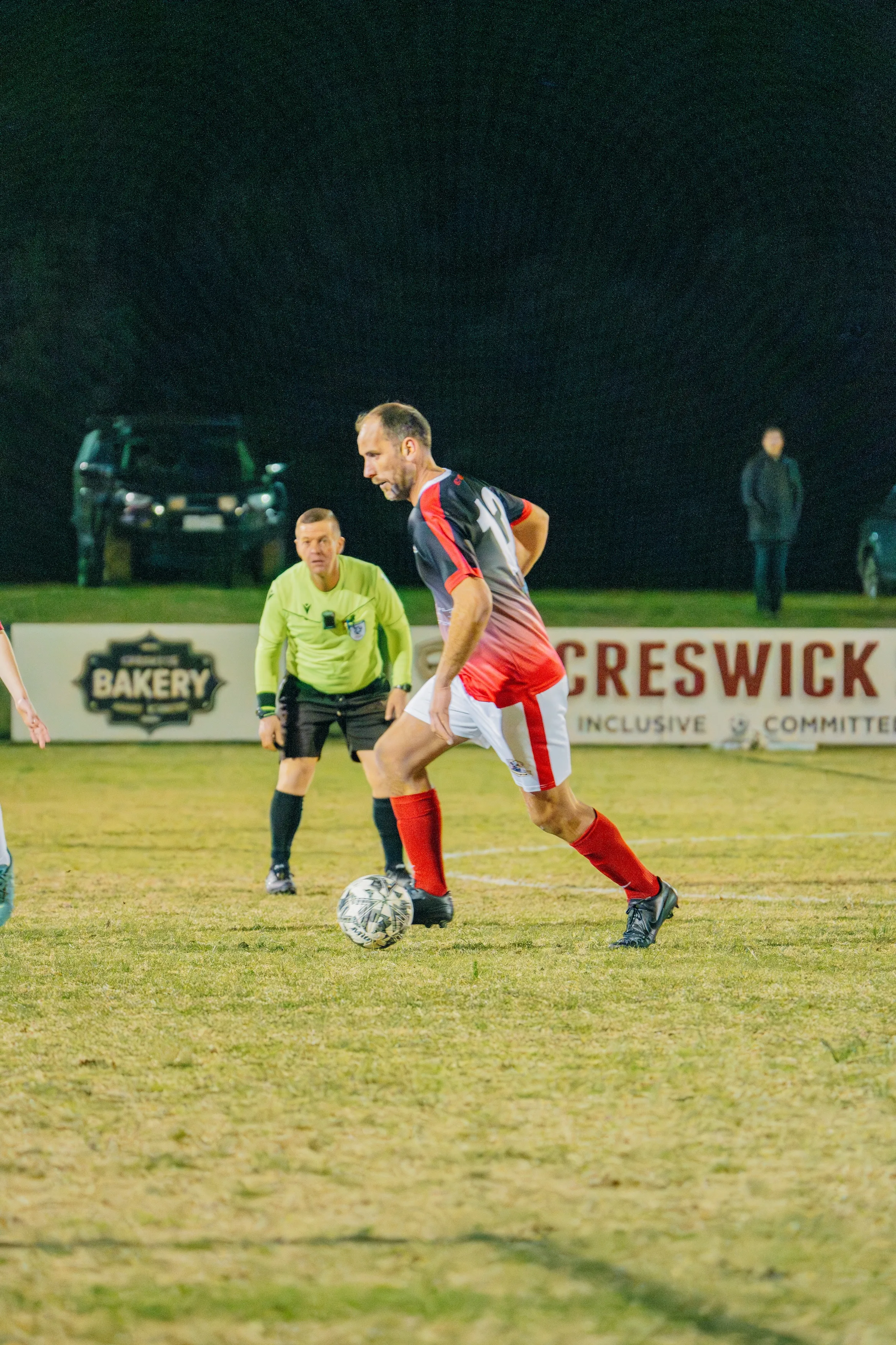 Soccer player in black and red jersey controlling the ball on a grass field with referee and a spectator in the background at night.