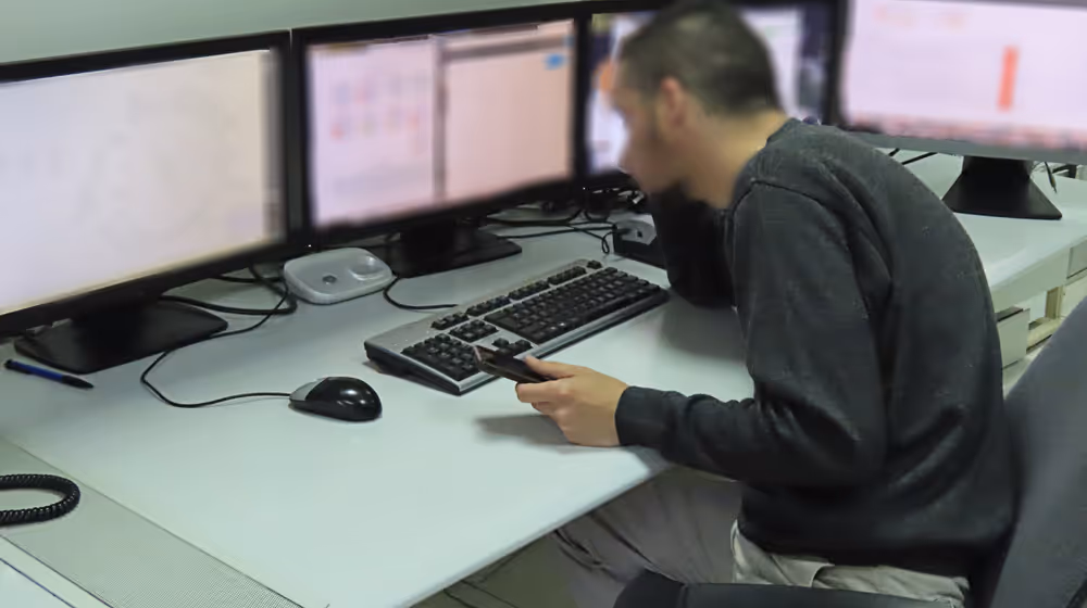 An individual in a control room setting, focused on multiple computer monitors displaying various graphs and interfaces. The person is seated, viewed from the side, and appears to be interacting with a device in hand, possibly managing or monitoring the event's digital aspects.