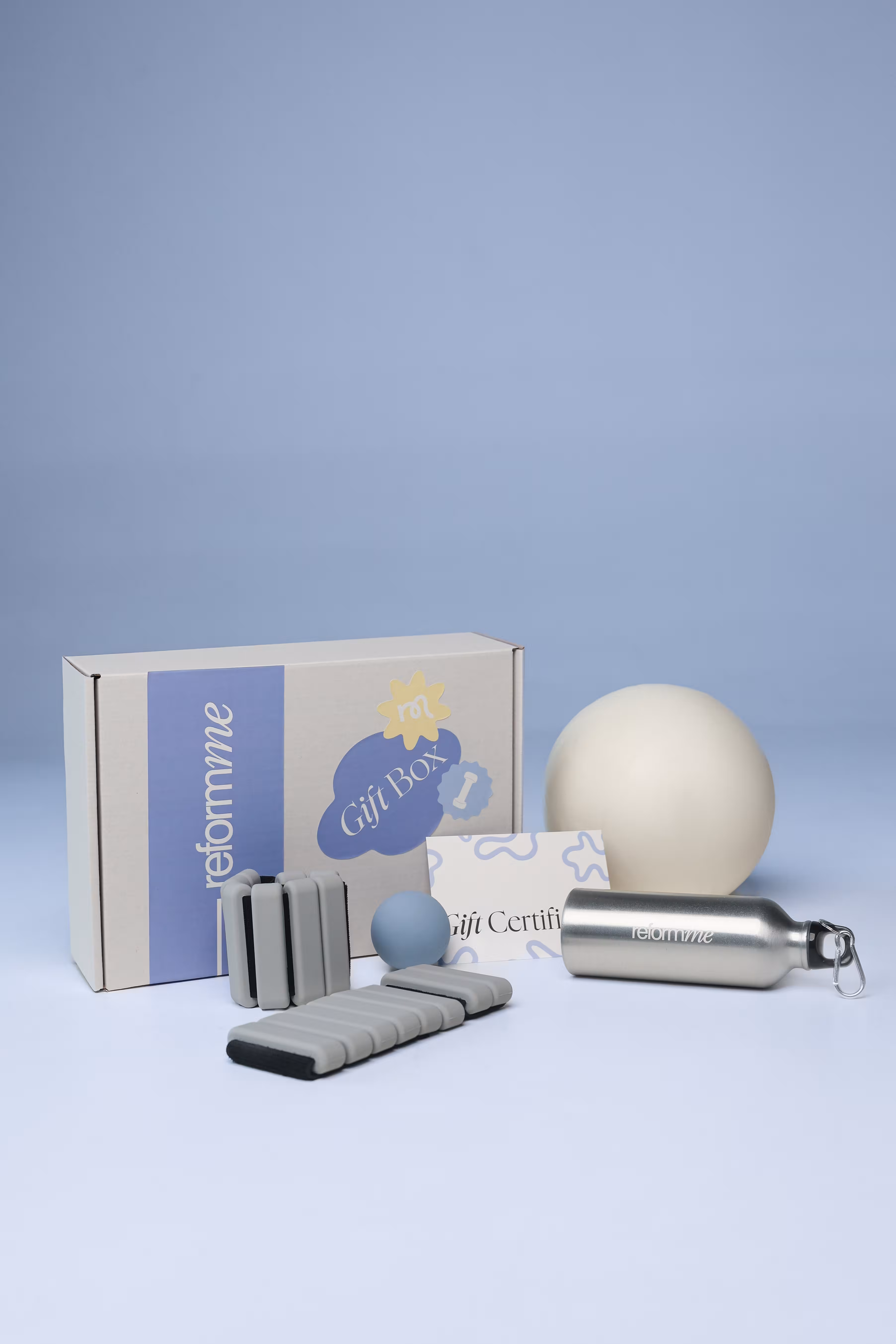 Reformme gift box set with a silver water bottle, two massage rollers, a small blue ball, a large white ball, and a gift certificate card on a blue background.