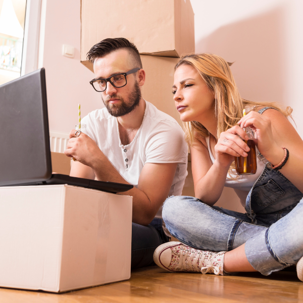 Couple sat on floor looking at a laptop surrounded by moving boxes