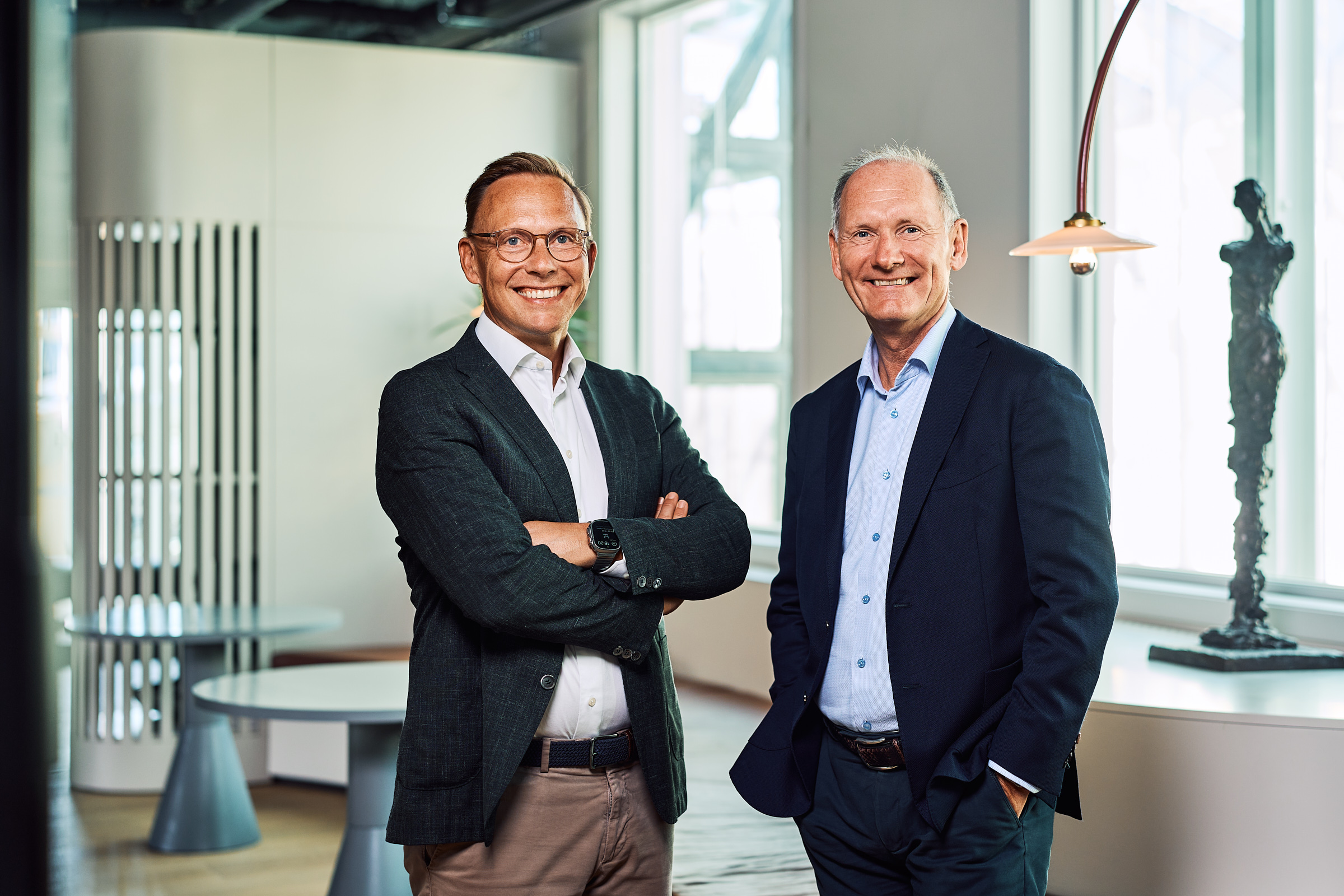 Two men in business attire smiling and standing in a bright modern office near windows and a sculpture.