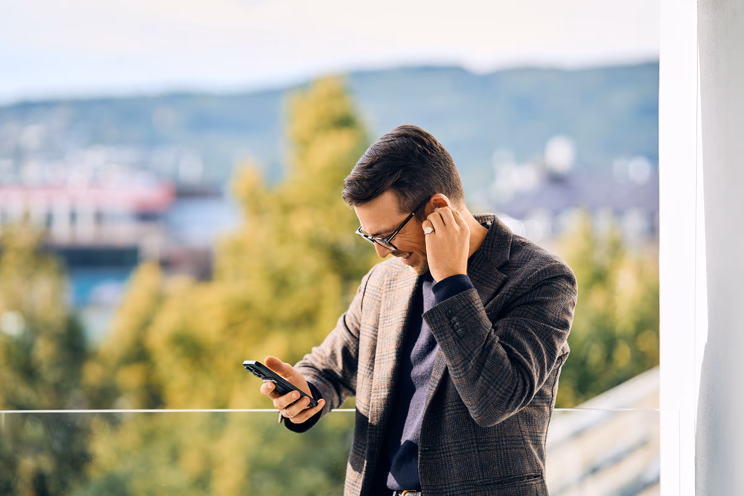 Smiling man adjusting wireless earbud while looking at smartphone on balcony with trees and hills in the background.