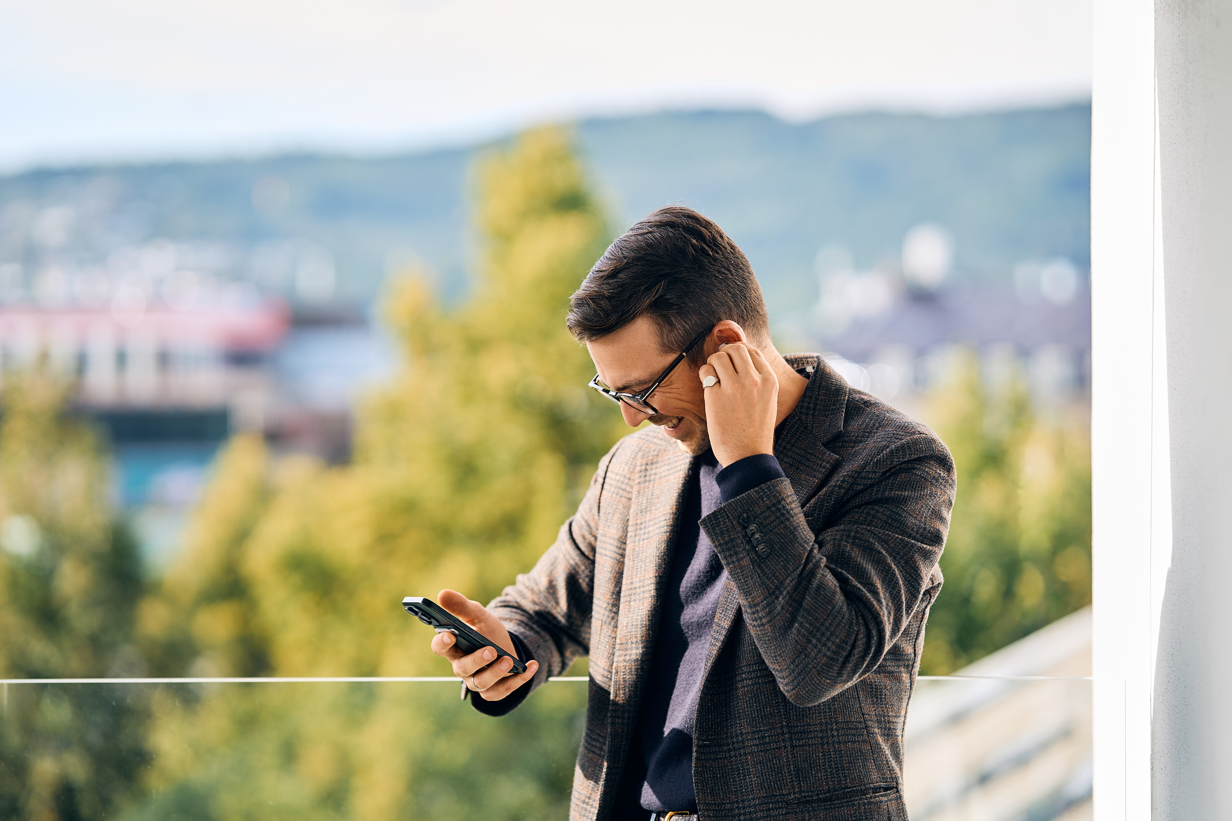 Smiling man adjusting wireless earbud while looking at smartphone on balcony with trees and hills in the background.