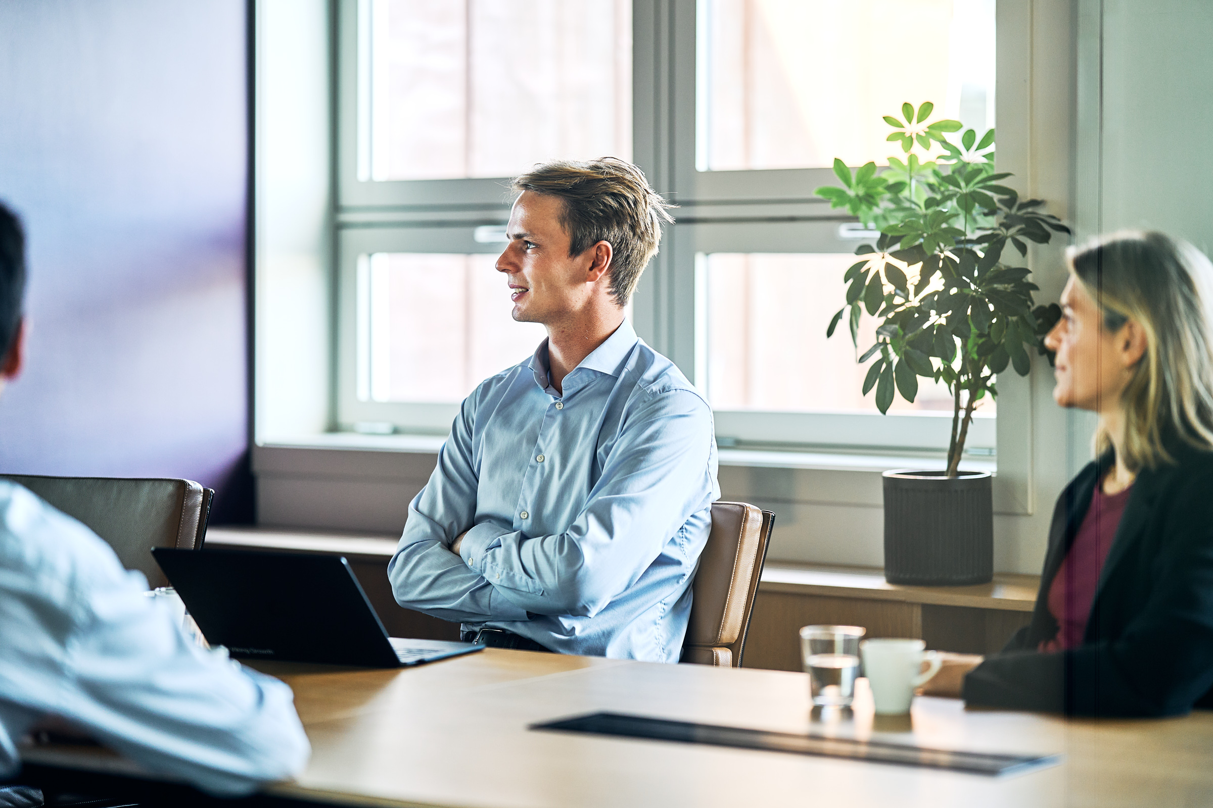 Young man in a blue shirt sitting at a conference table with a laptop, engaged in discussion with others in a bright meeting room.