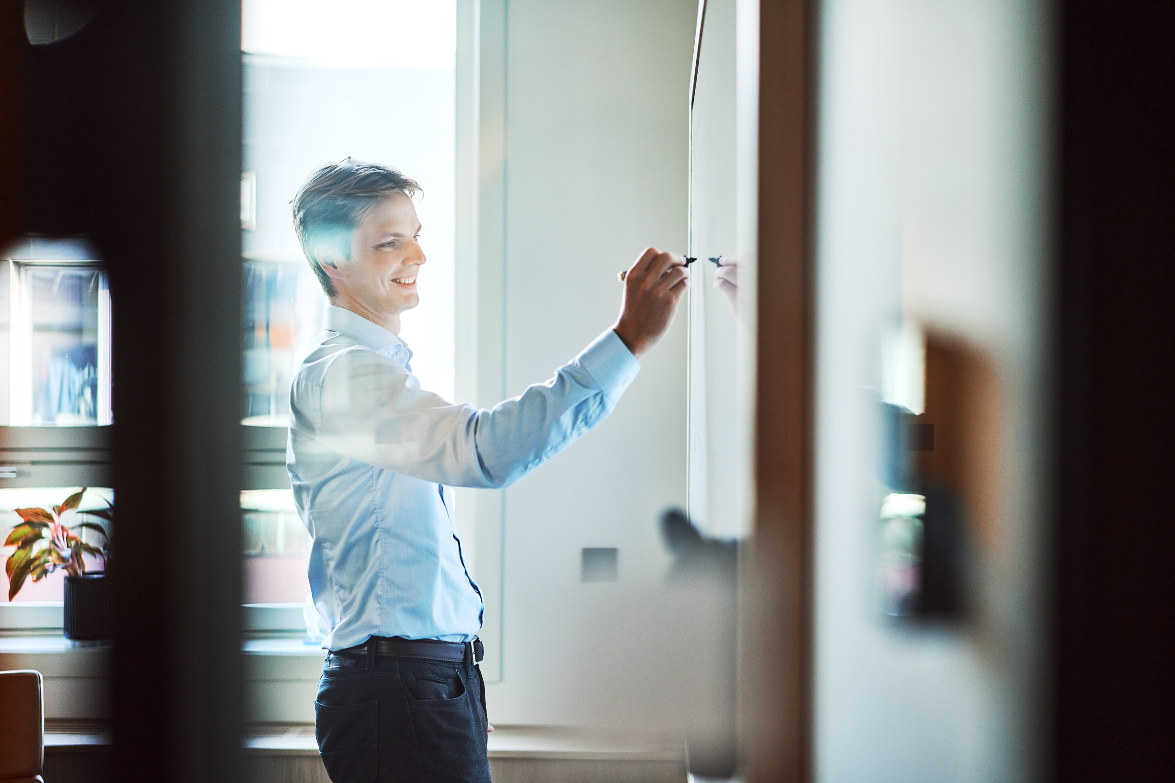 Smiling man in light blue shirt writing on a whiteboard in a bright office.