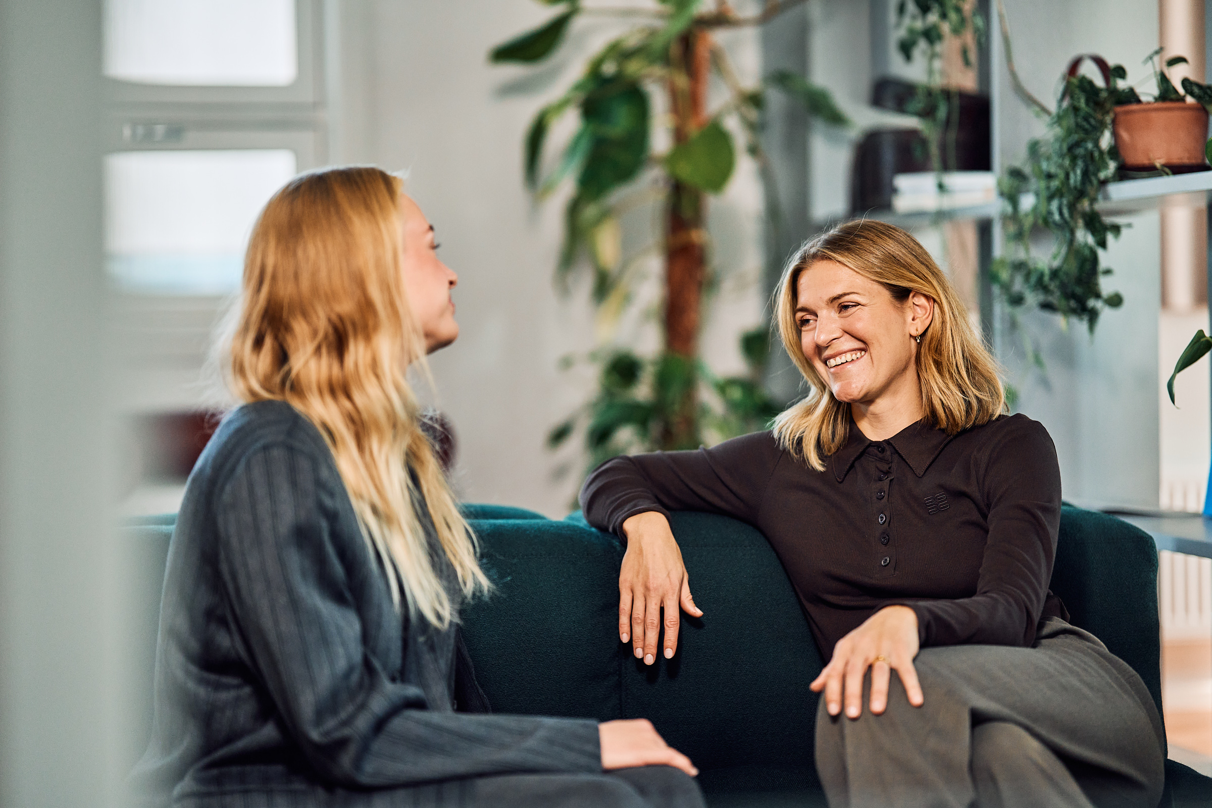 Two women smiling and having a conversation while sitting on a dark green sofa in a room with plants.
