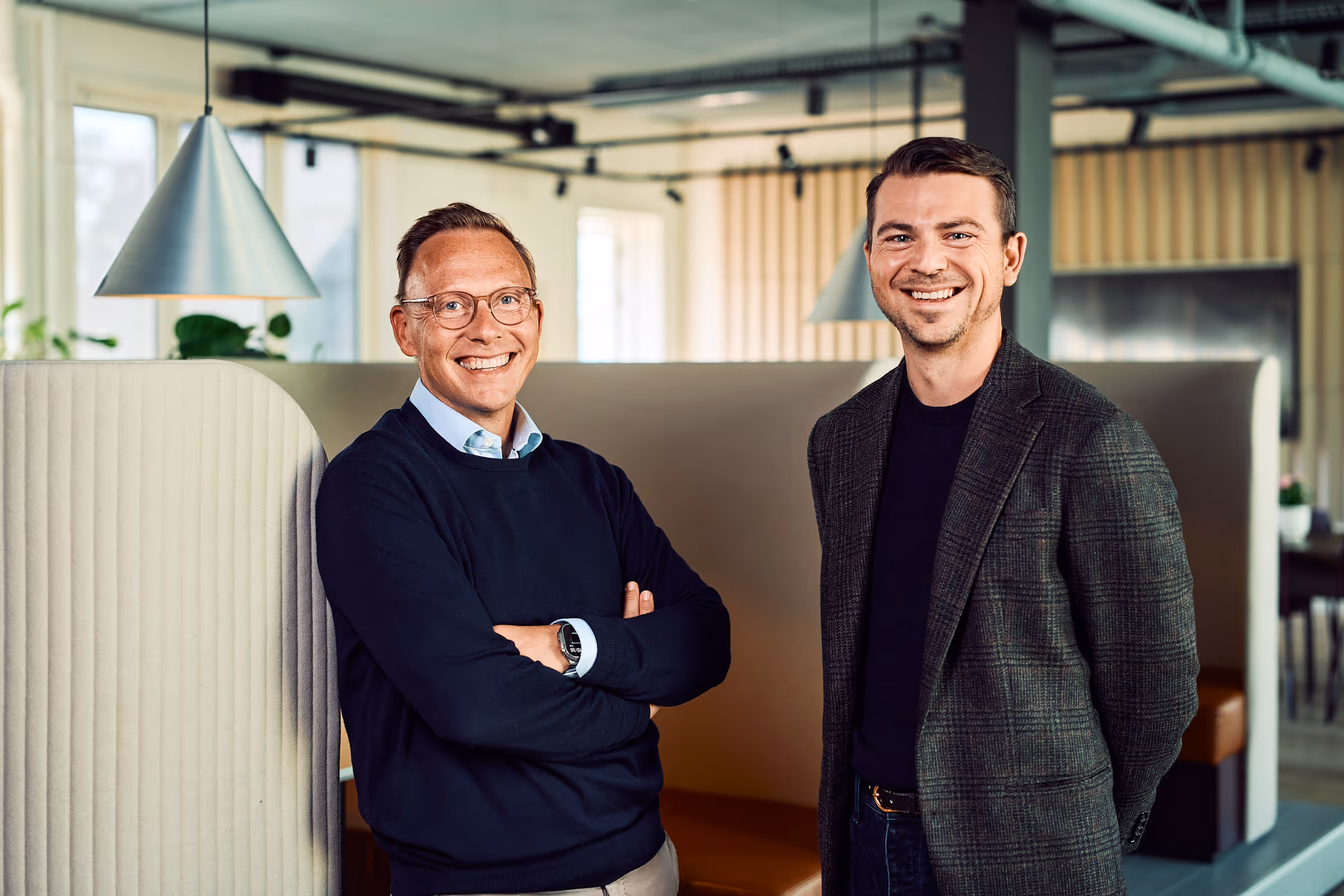 Two men smiling and standing side by side in a modern office space with beige partitions and hanging lights.