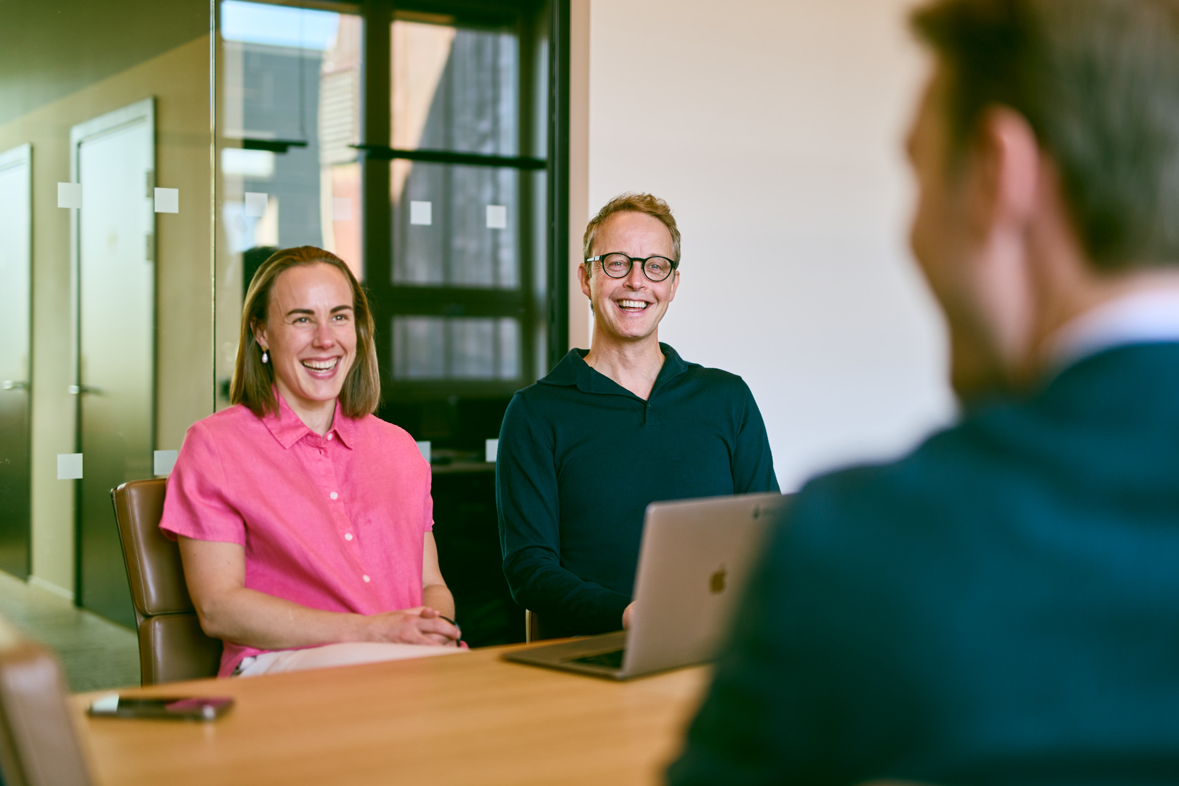 Two people smiling and sitting at a table with a laptop during a meeting, viewed from behind a third person.