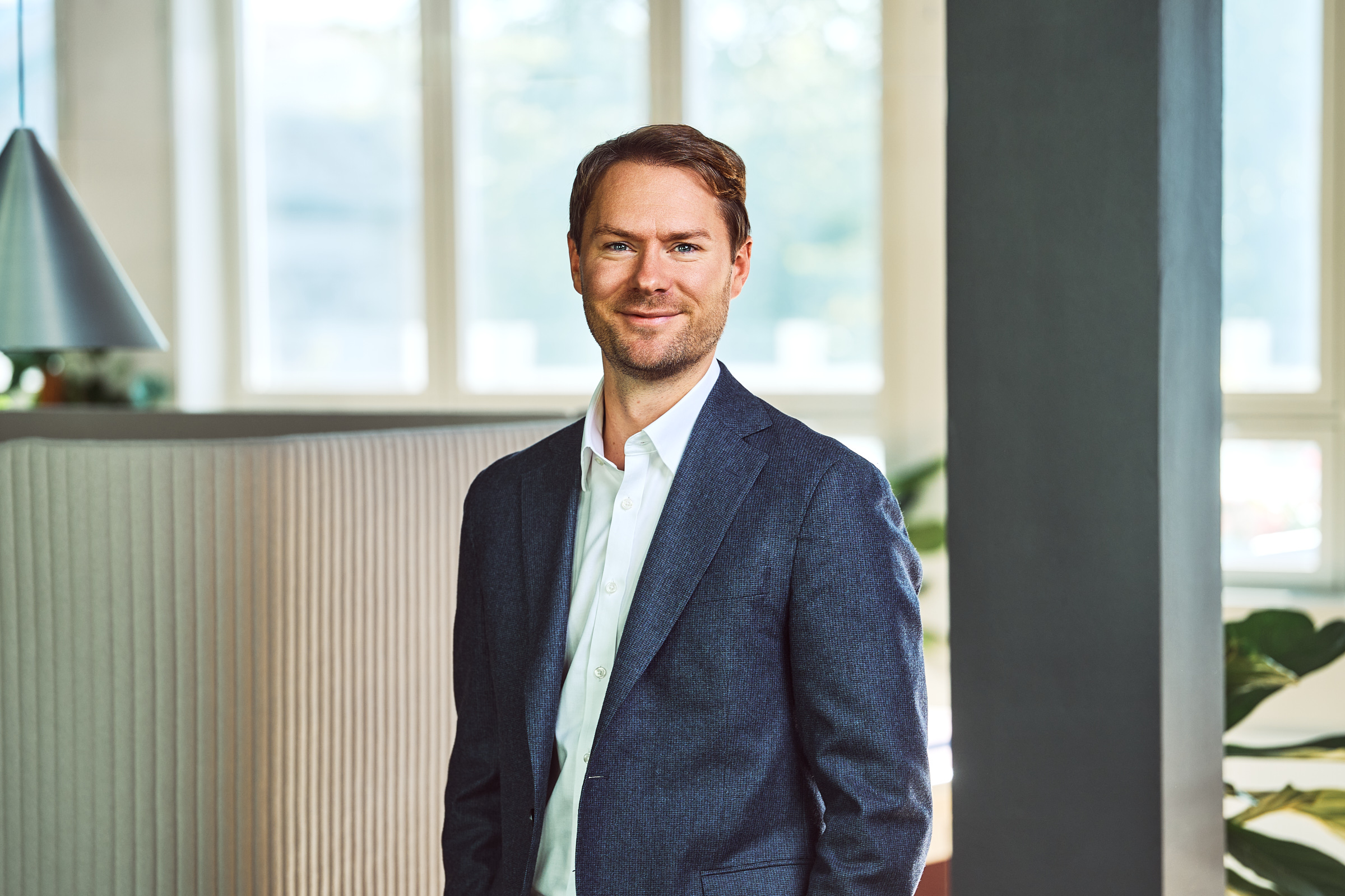 Smiling man in a navy blazer and white shirt standing indoors with modern office background.