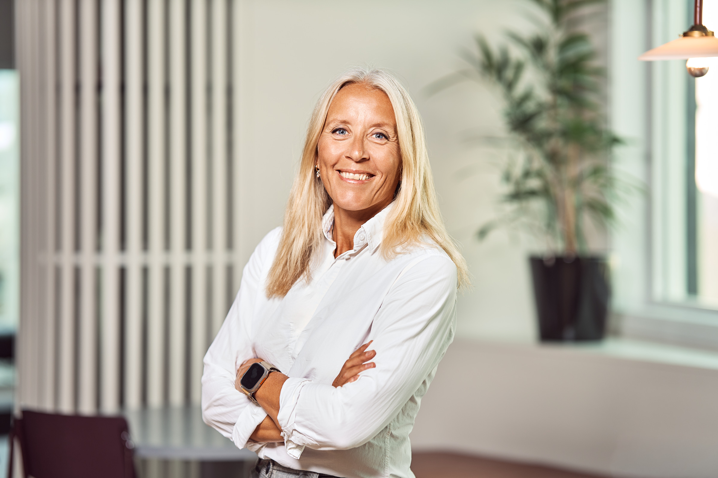 Smiling woman with long blonde hair, wearing a white shirt and a smartwatch, standing with arms crossed in a bright office space.