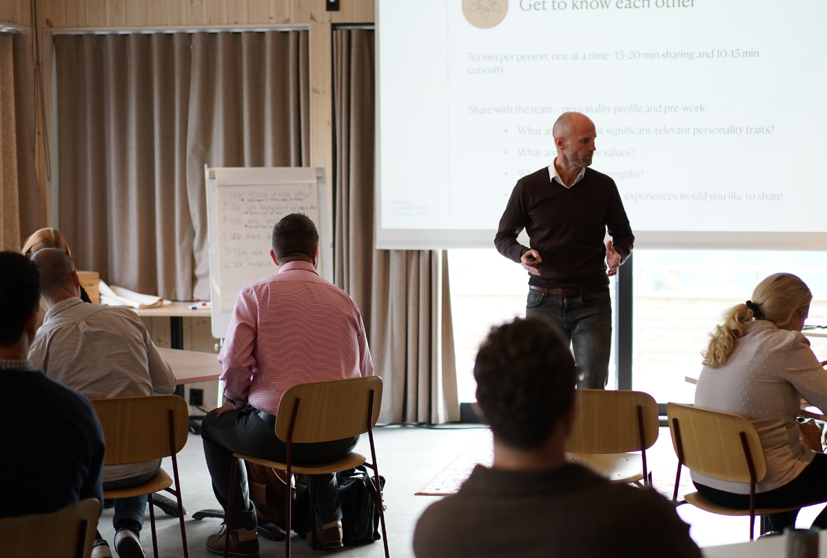 Man standing and speaking in front of a seated audience in a meeting room with a presentation slide and flip chart in the background.