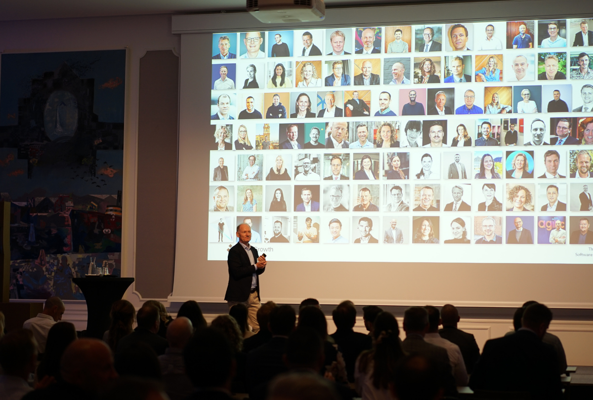 Man in a suit speaking on stage in front of an audience with a large screen displaying a collage of headshots.