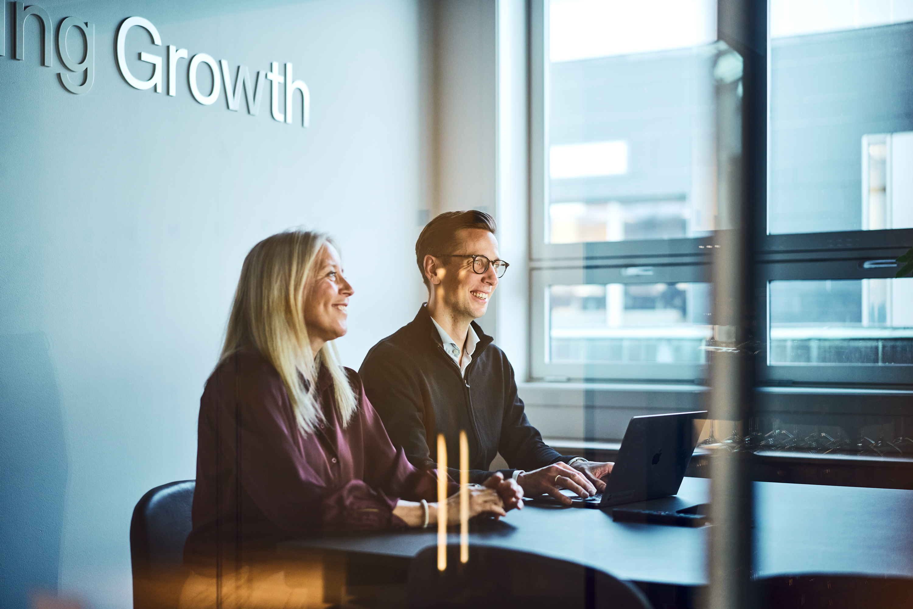 Two colleagues sitting at a table in a bright office, smiling and working on a laptop near a window.