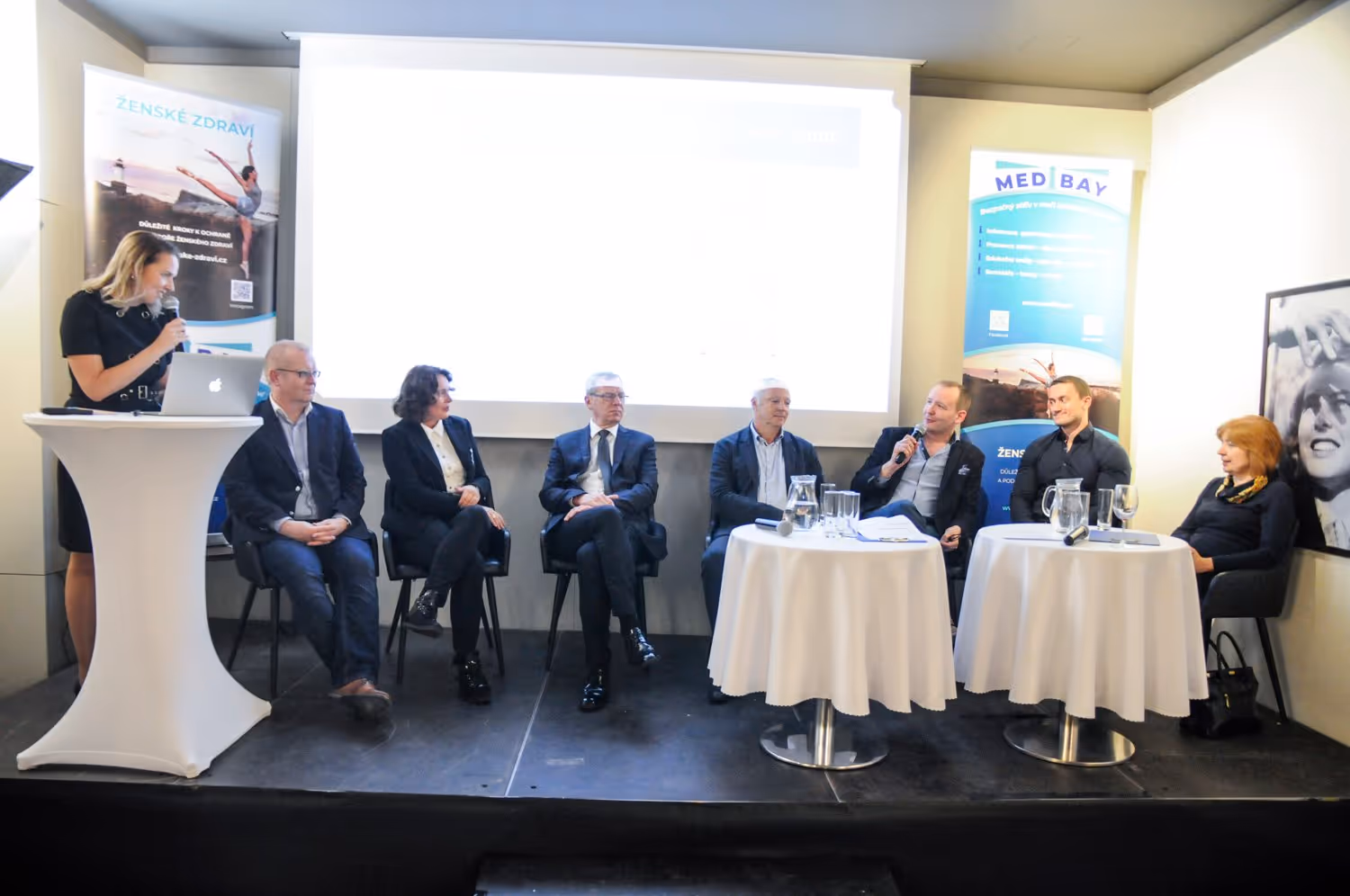 Řečníci sedící na židlích na pódiu.