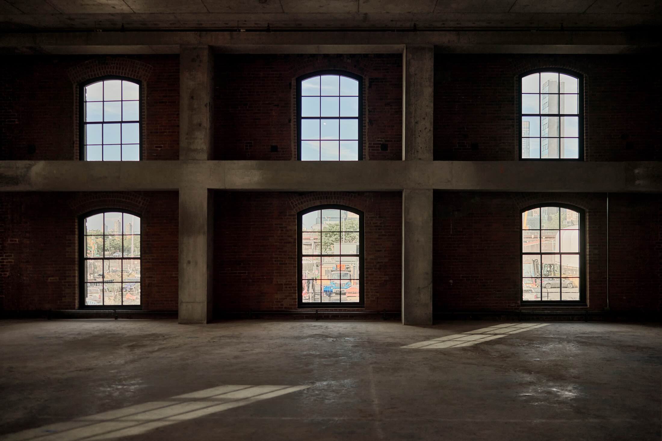 Interior view of the dimly lit, unfinished event hall inside The Mallory NYC showing raw concrete floors, exposed red brick walls, and double-stacked arched windows with natural light casting geometric shadows.
