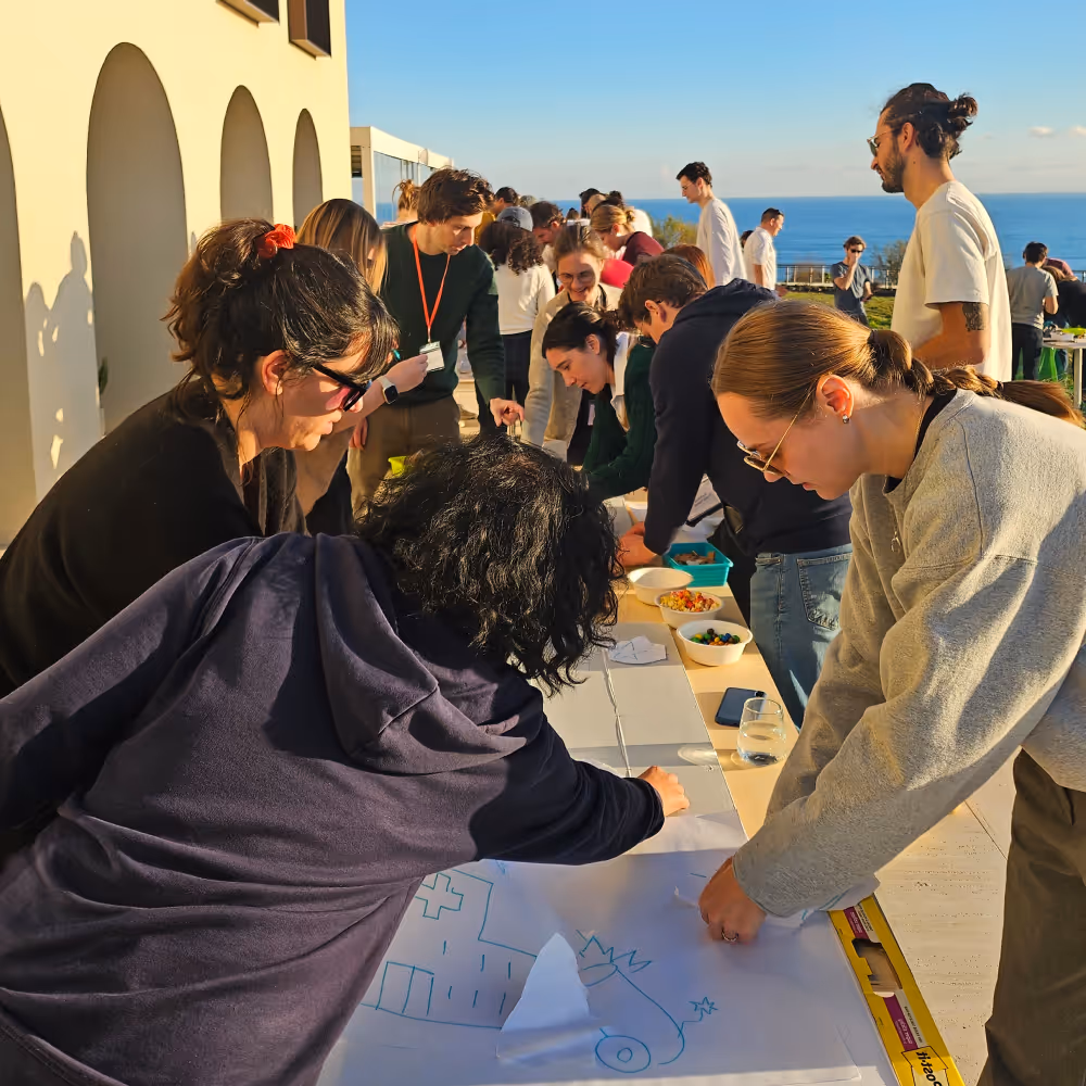 Group of people gathered around tables outdoors near a coastal area, engaging in an activity under clear blue sky.