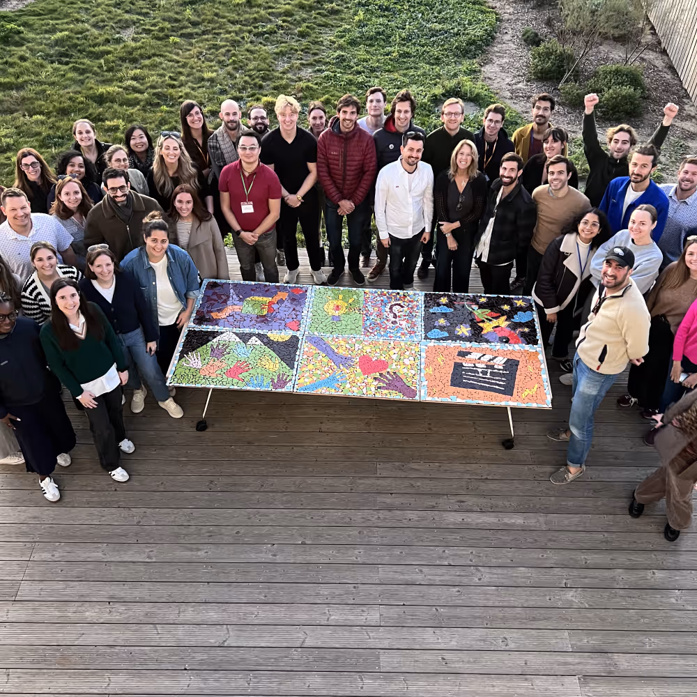 Group of people standing around a table displaying colorful mosaic artwork on a wooden deck with grass in the background.