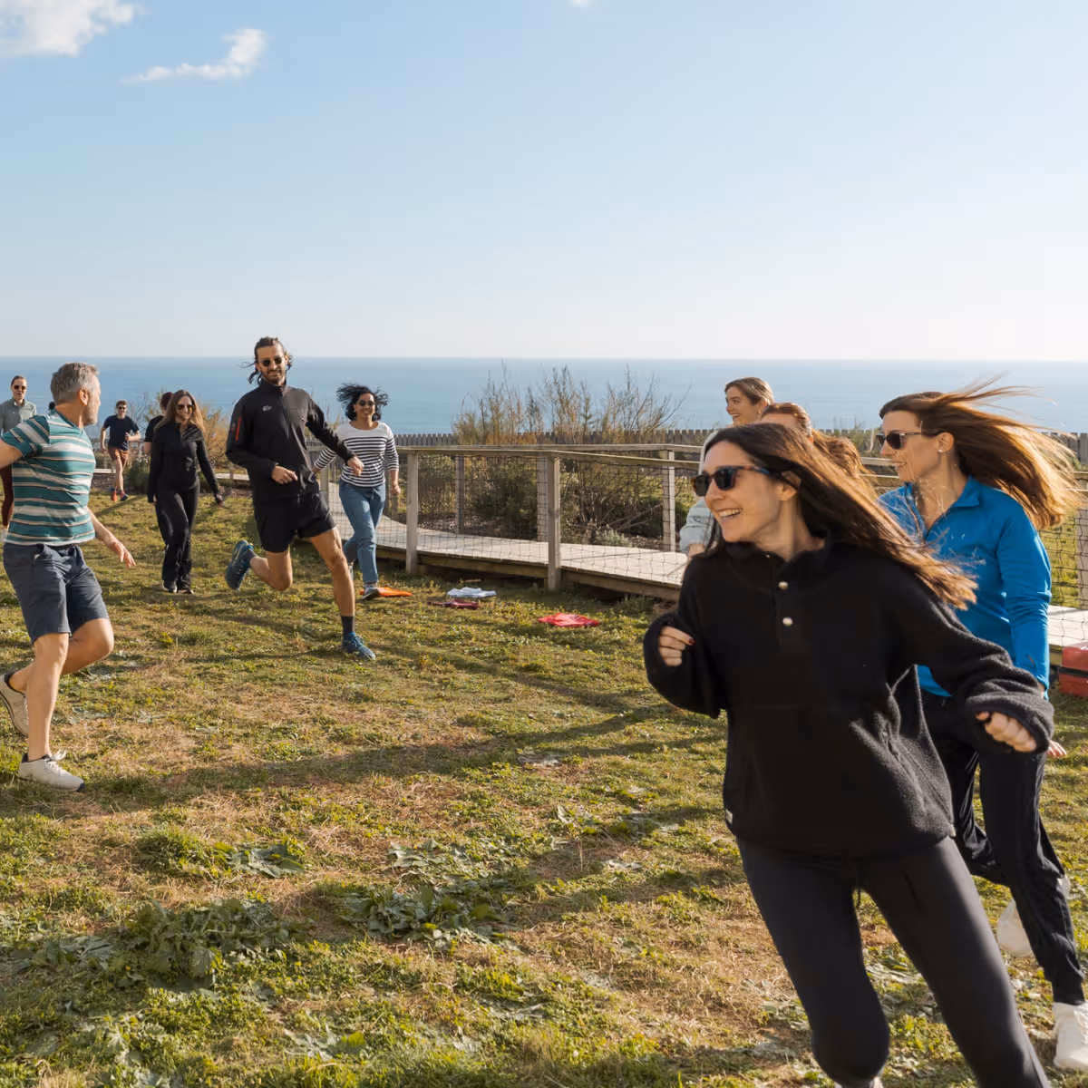 Group of people running and playing outdoors on grass near a wooden walkway with ocean in the background.