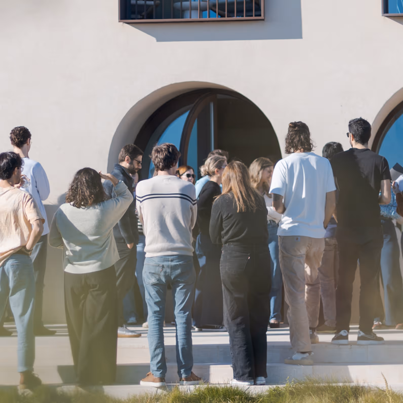 Group of people standing outside near a building with large arched doorways, experiencing sunlight.