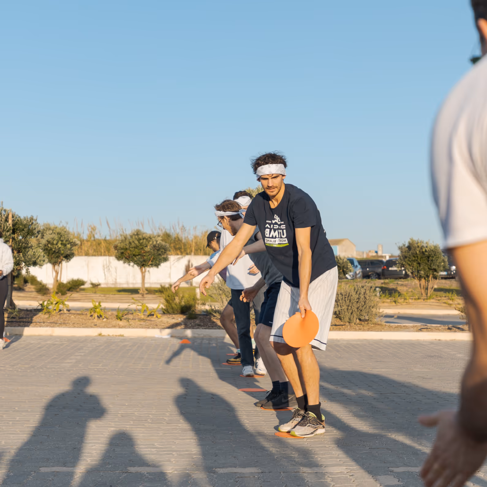 Group of young men standing in a line outdoors, each holding an orange flying disc, preparing to throw it.