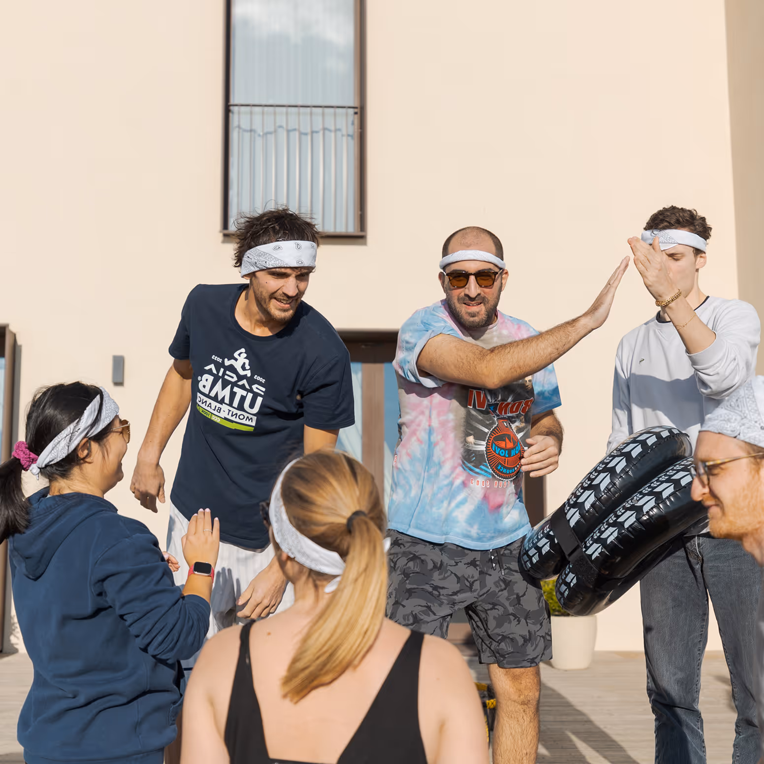Group of six young adults wearing headbands, outdoors, giving high fives and holding inflatable tires during a daytime activity.