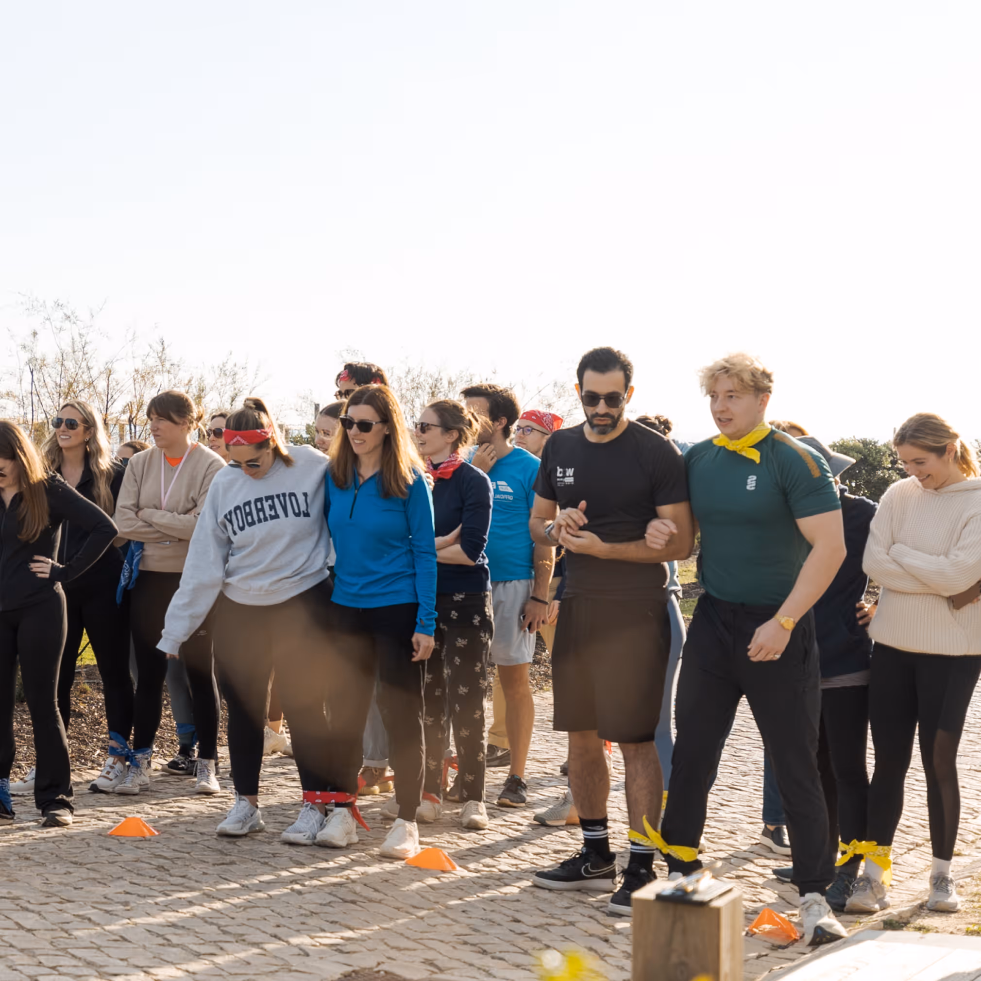 Group of young adults outdoors participating in a three-legged race with their ankles tied together by colored bands.