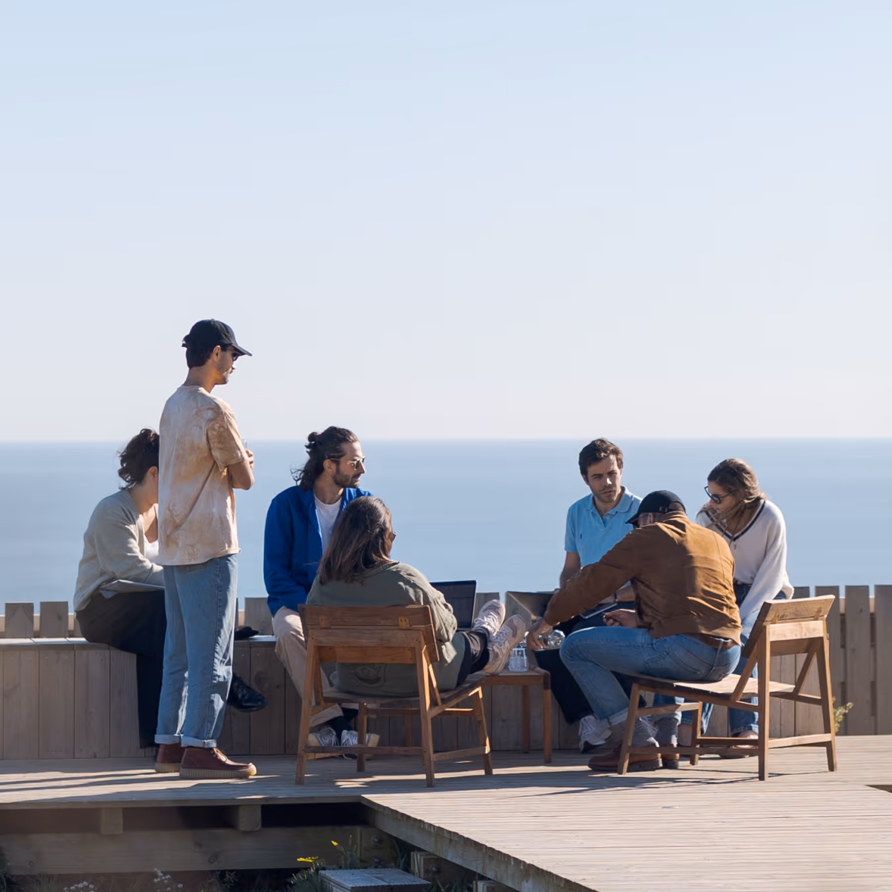 Group of six people sitting and standing on a wooden deck with laptops, overlooking the ocean on a clear day.
