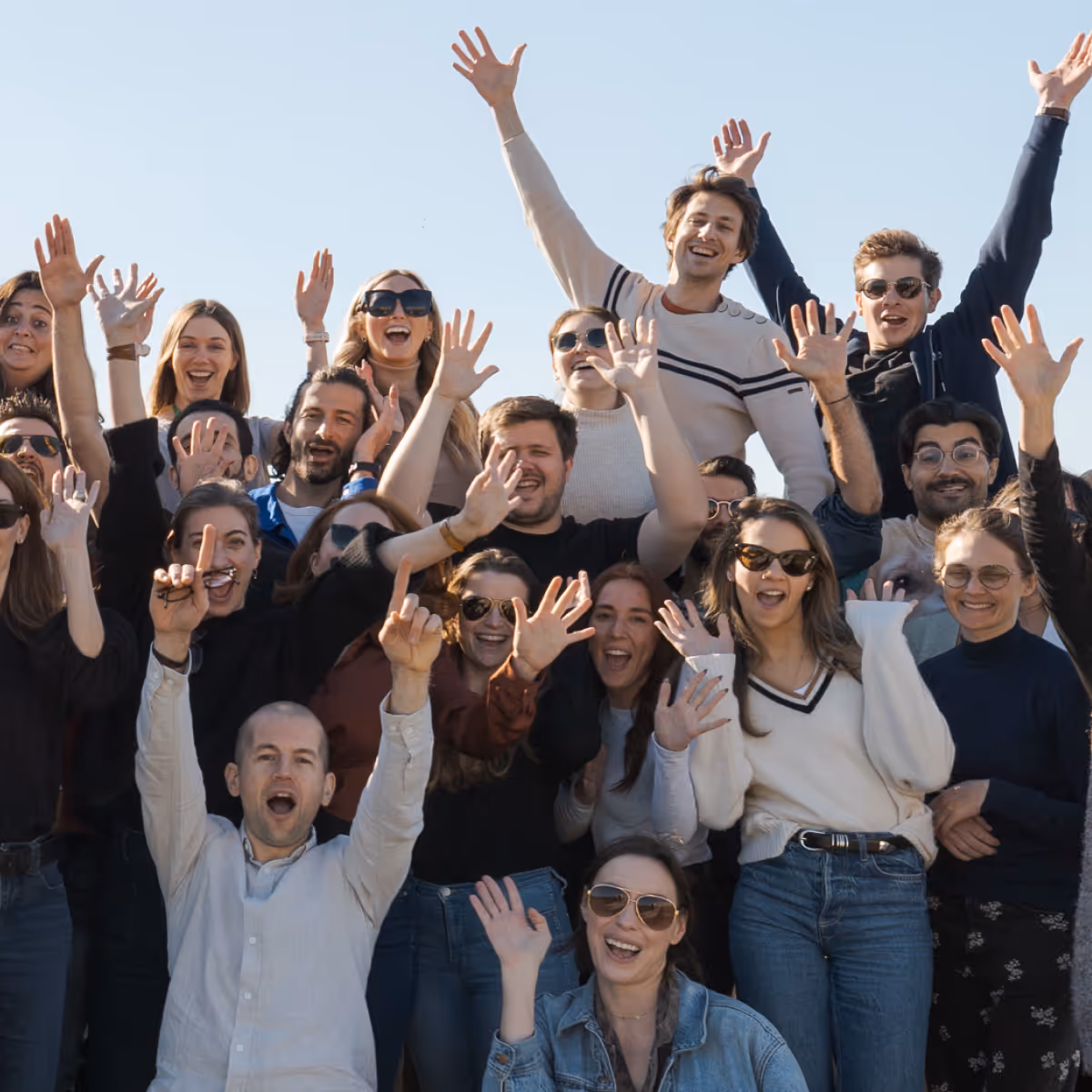 Group of diverse young adults joyfully raising their hands and smiling outdoors under clear sky.