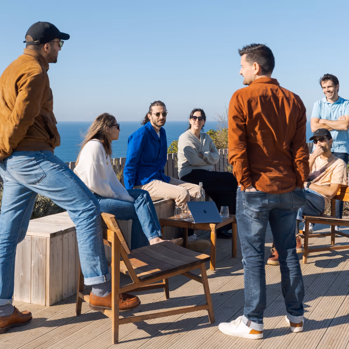 Group of seven adults casually gathered outdoors on a wooden deck overlooking the ocean, some seated and others standing, engaging in conversation on a sunny day.