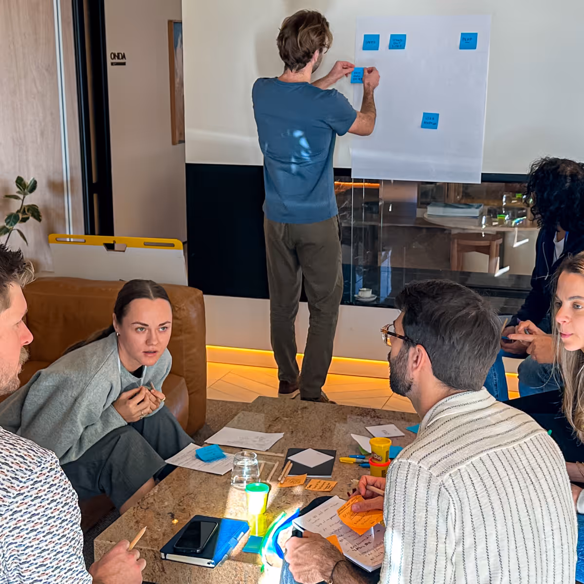 Group of five people in a meeting room brainstorming with sticky notes on a whiteboard and papers on a table.