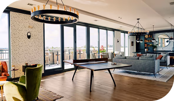 Spacious modern living room with hardwood flooring, featuring a ping pong table, green armchair, and large windows overlooking a balcony. Contemporary lighting fixtures and a cozy seating area complete the stylish interior design.